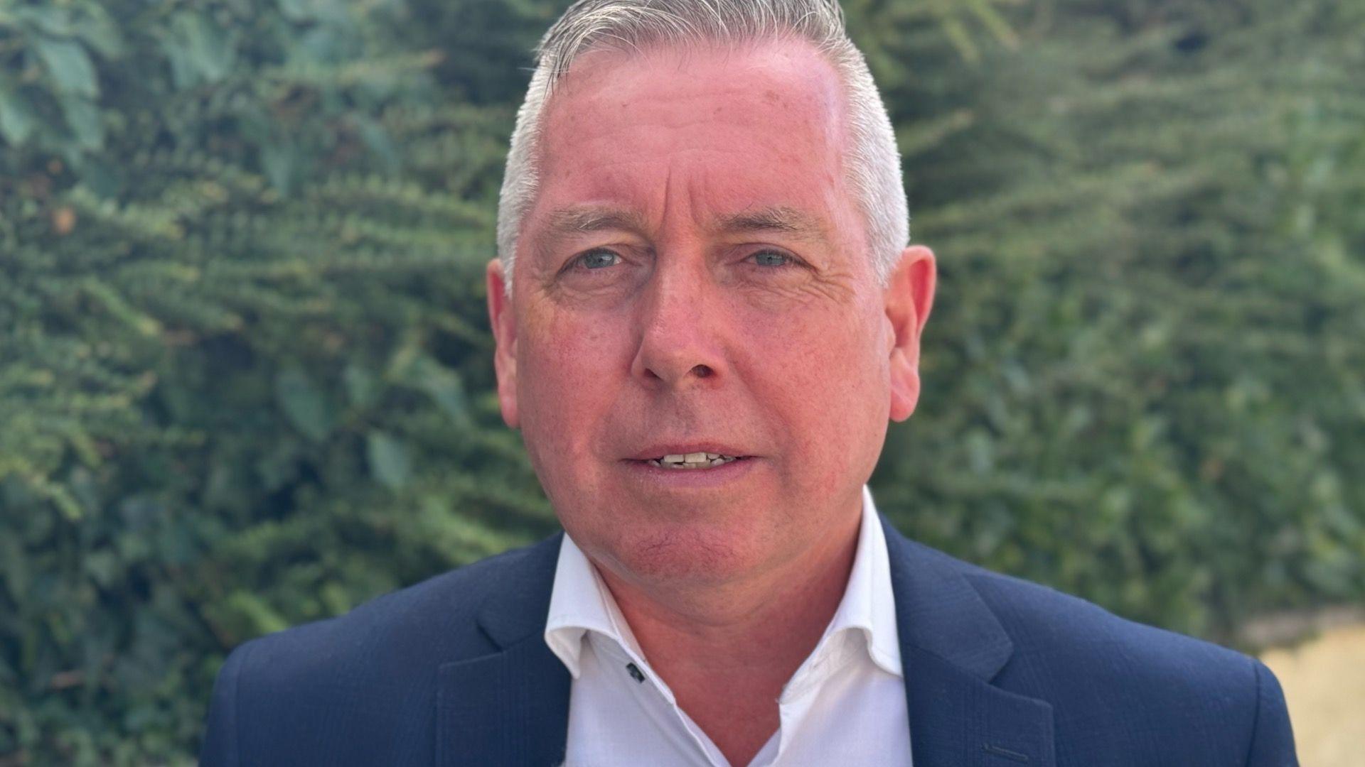 A man with short grey hair, a navy jacket and a white shirt. 
