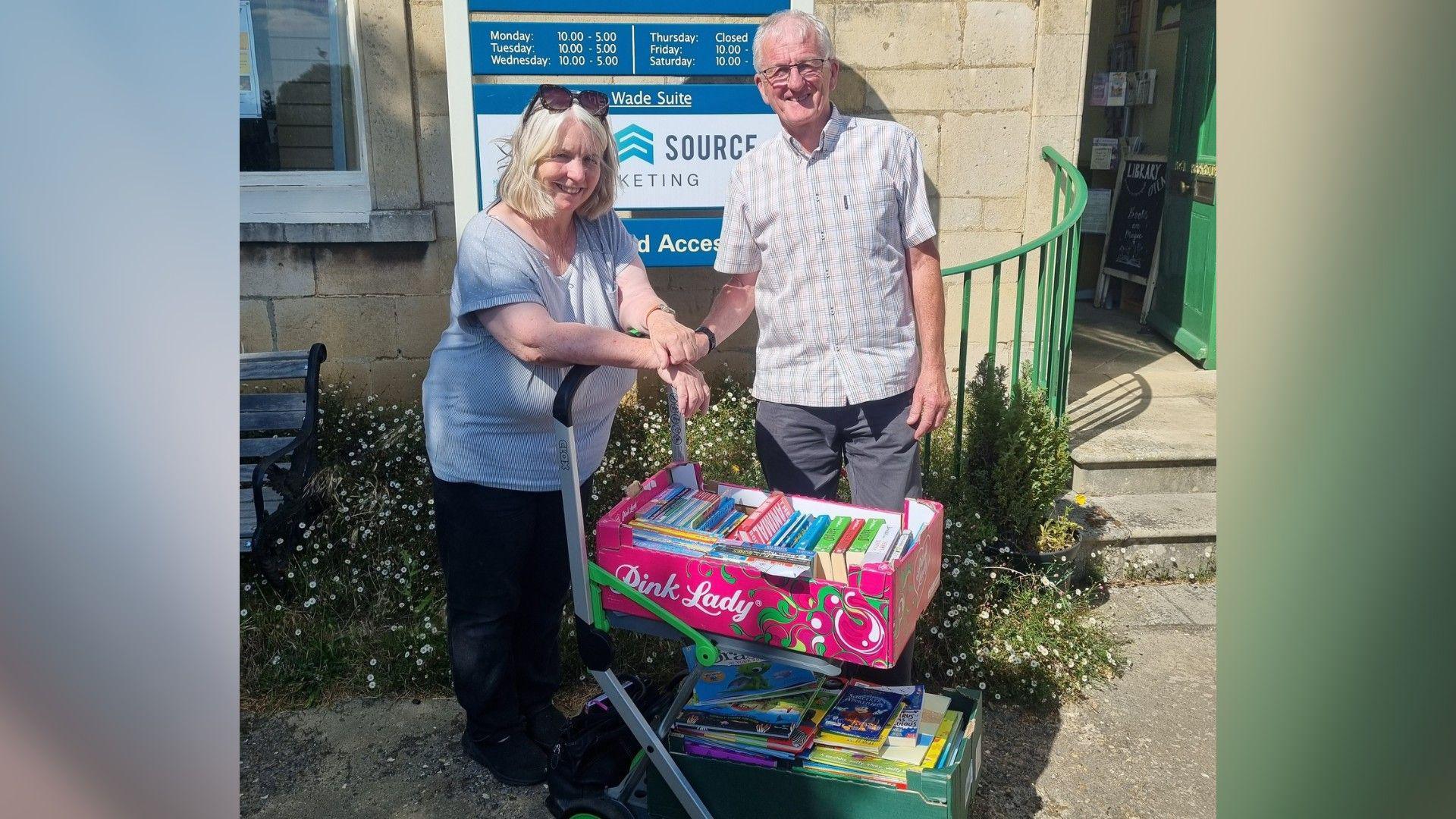 Volunteers from Deeping Library- a lady wearing a grey T-shirt and Black trousers and a man wearing a checked shirt standing behind a trolley with two crates of books