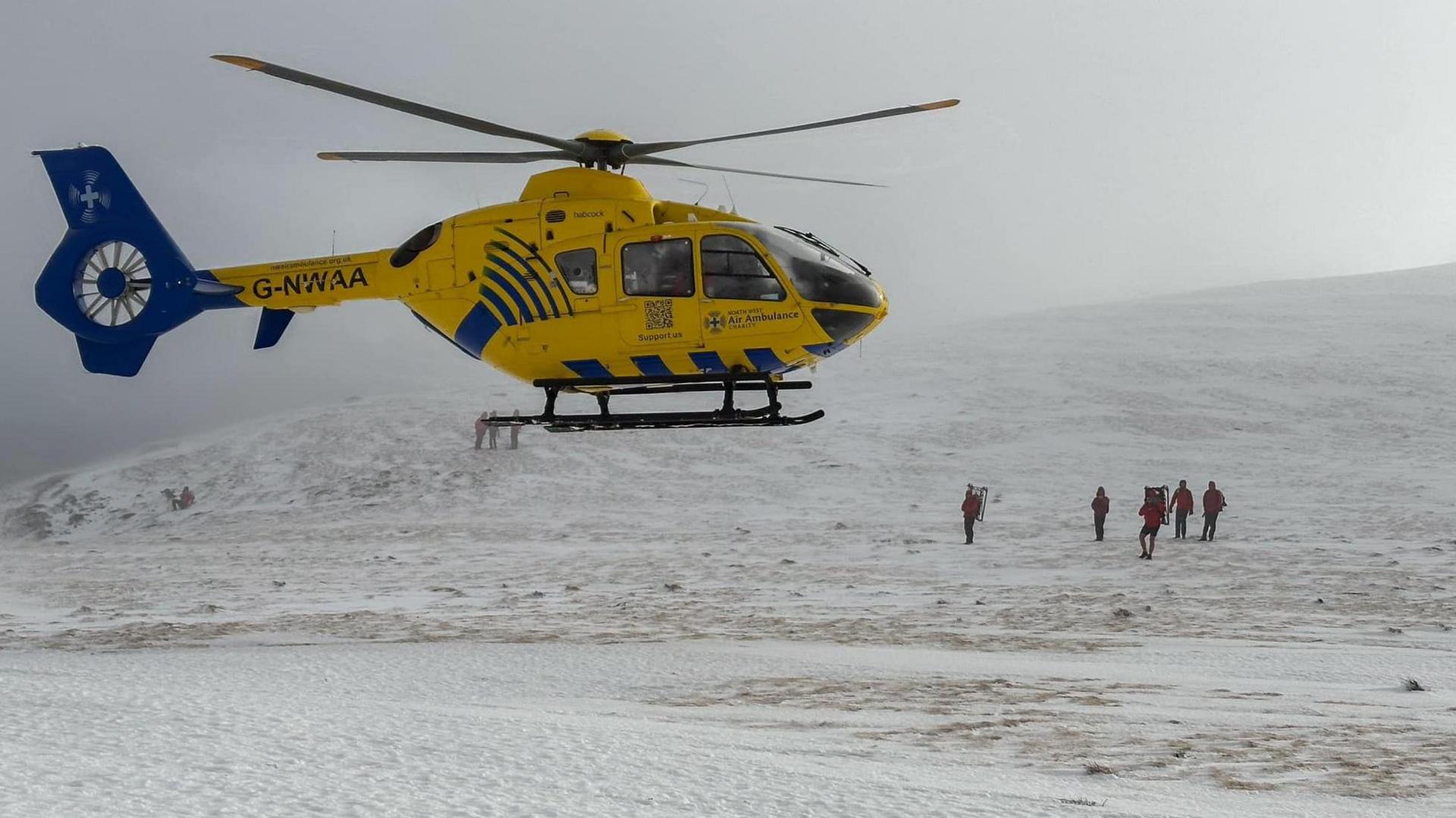 A team of Keswick Mountain Rescue volunteers, who are all wearing red waterproofs and large rucksacks, stand on top of the summit of Blencathra. There is a yellow helicopter hovering above them.