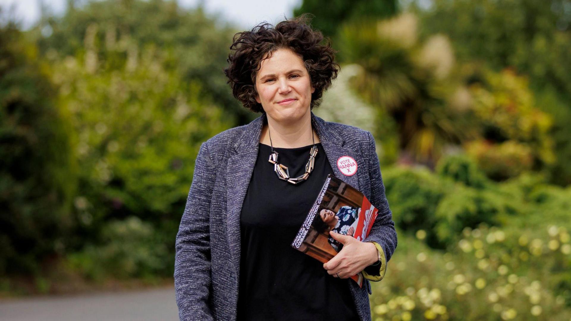 Claire Hanna standing in outside in a grey jacket and black top holding some election leaflets