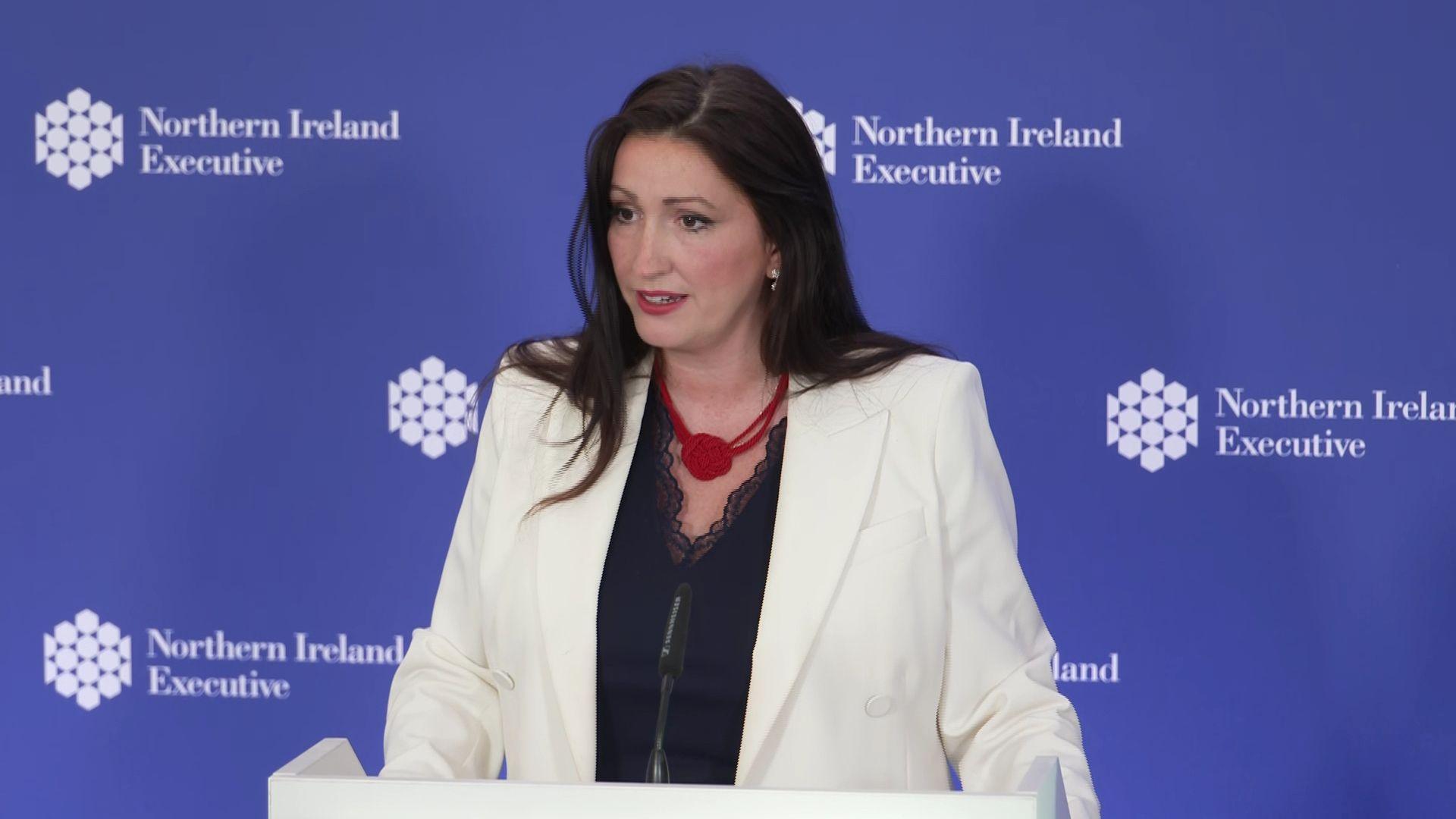 Emma Little-Pengelly stands at a mic. She wears a blue top and white blazer. She has brown hair.