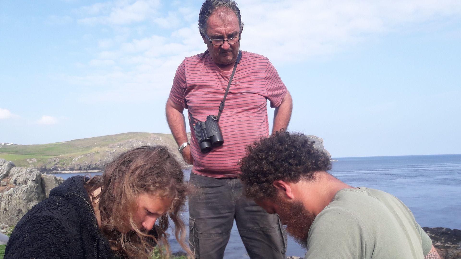 Aron Sapsford wearing a pink T-shirt and binoculars standing above a younger man and woman who are sitting down ringing birds.