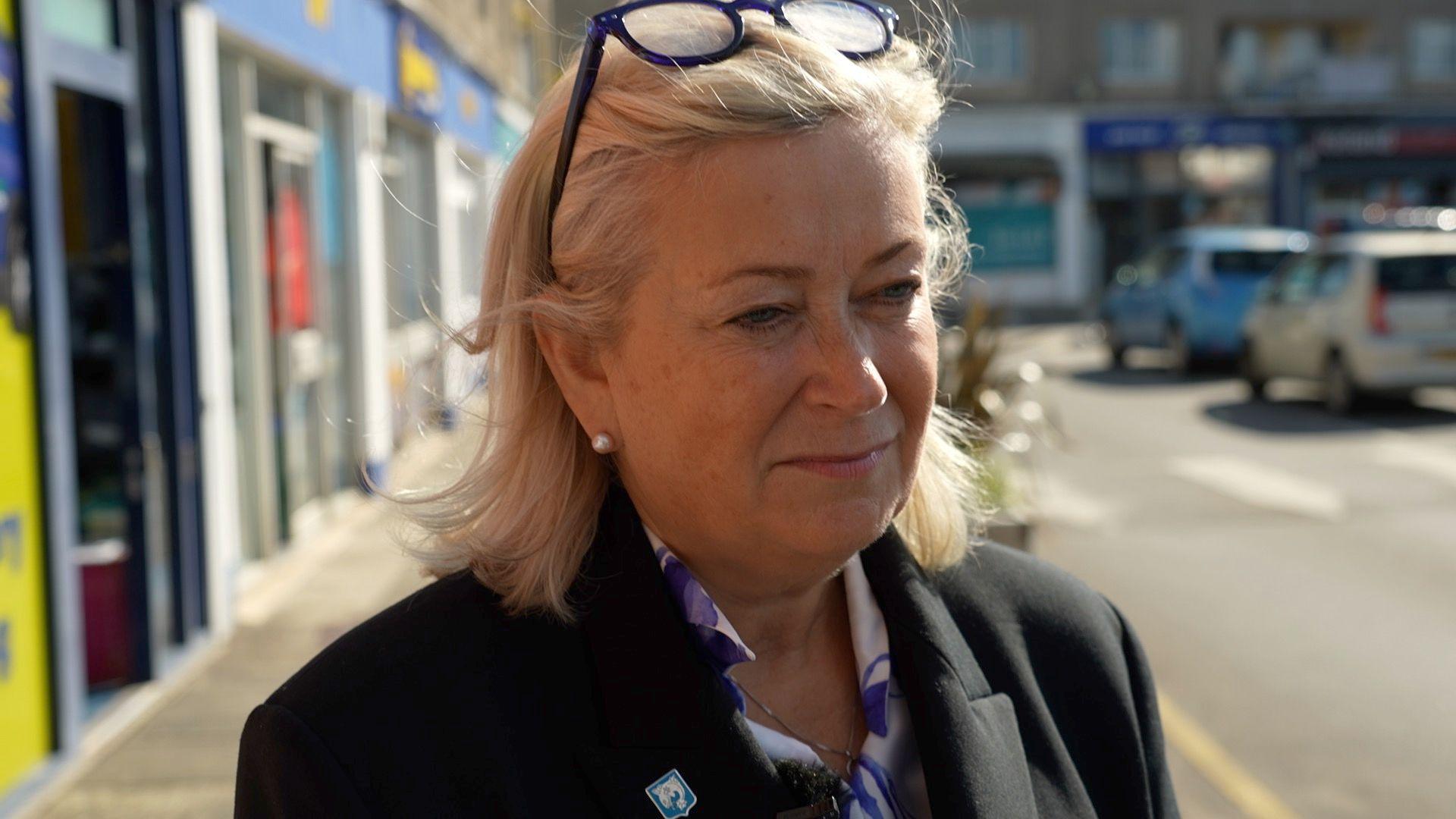 A woman with blonde hair, dark glasses and a black suit jacket and blue and white shirt on a street in Jersey. There are cars and shops behind her.