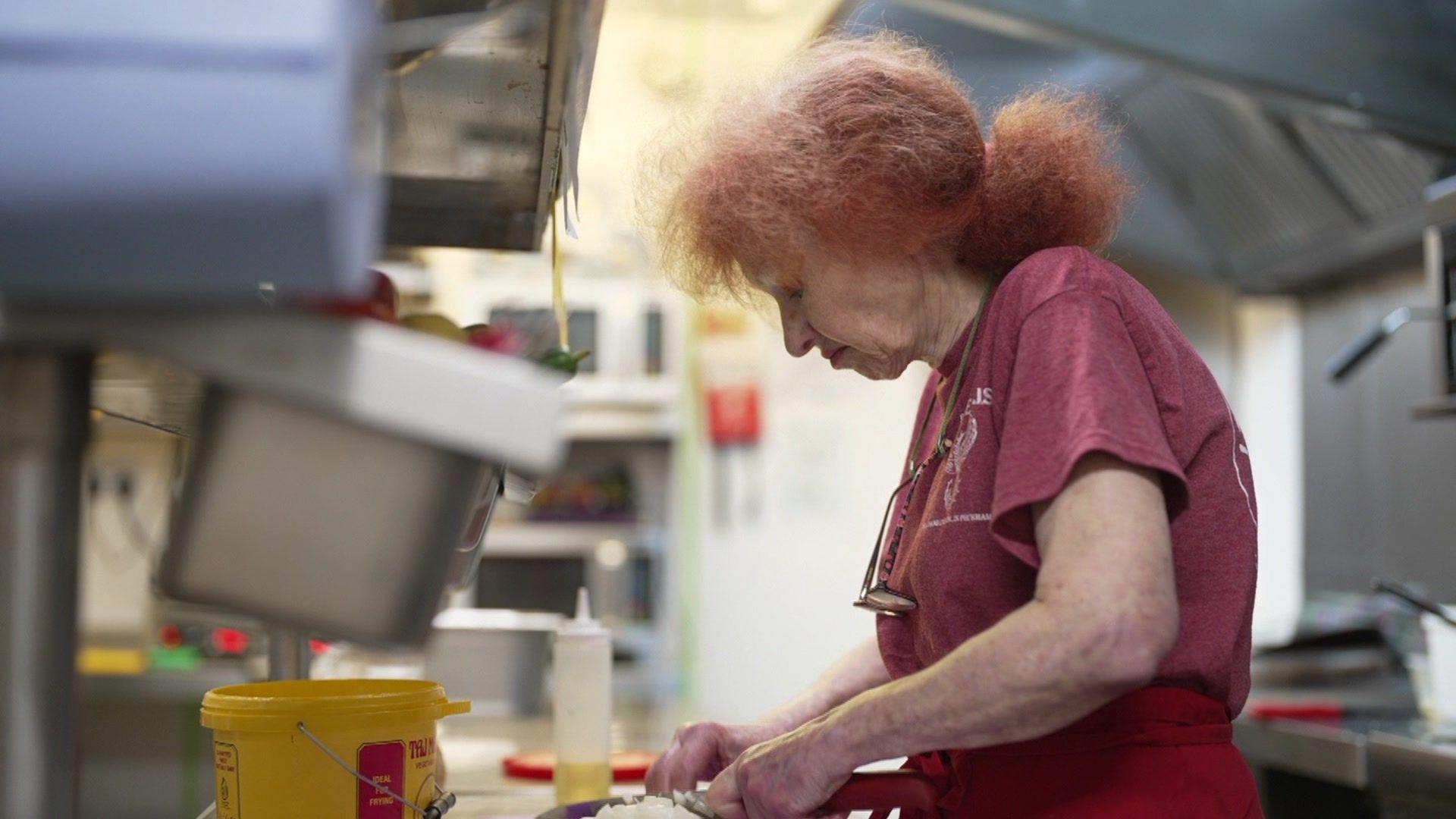 Sally butcher is seen cutting onions in her restaurant kitchen. She is wearing a red T-shirt and apron. 