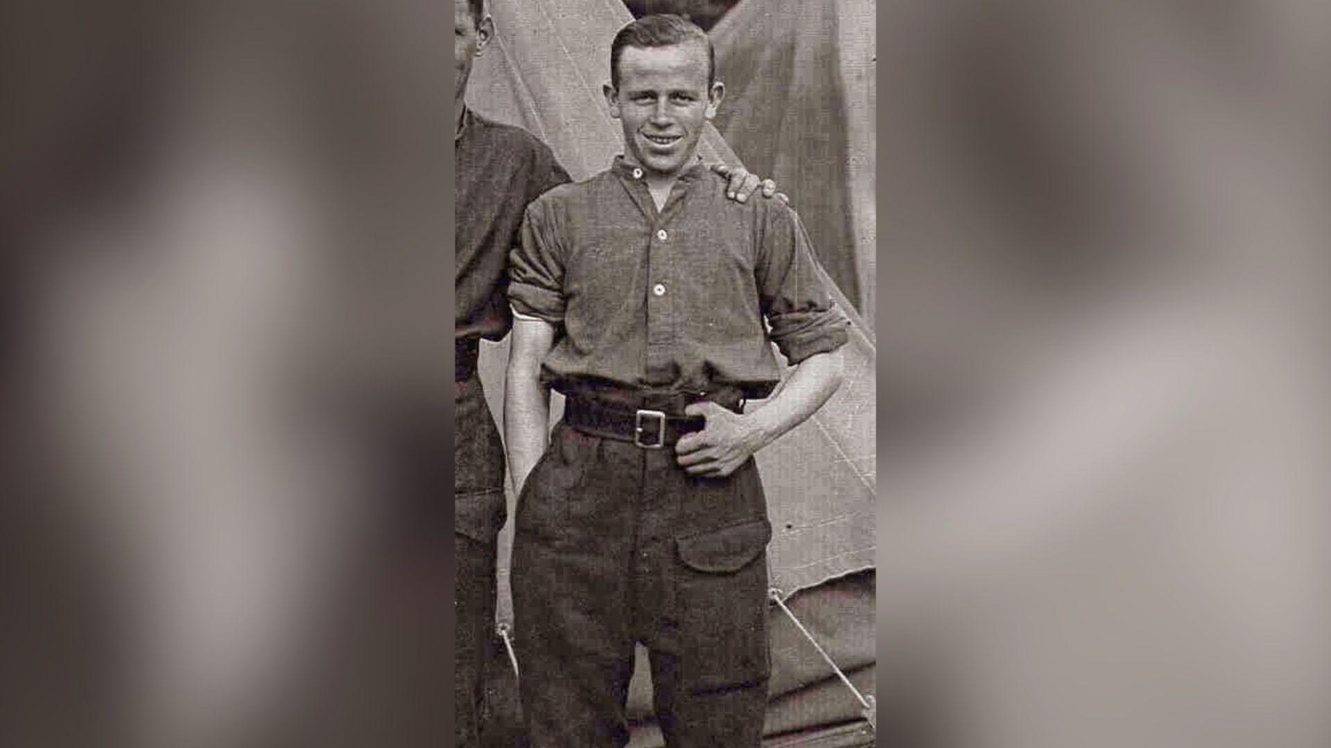 A man in an Army uniform standing in front of a tent. He has brown hair and is smiling at the camera. There is an arm around his shoulders.