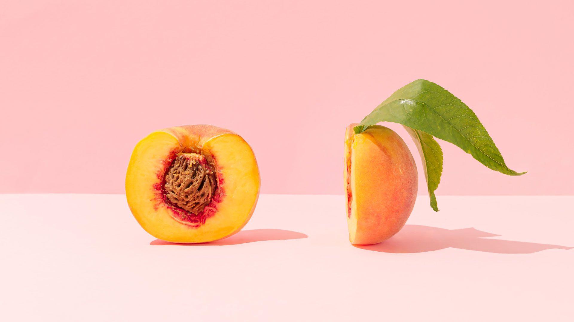 Peach cut in half, the left side is facing the front with the stone in it, the other half is on it's side with the leaf coming out of the top. The peach sits on a pale pink background. 