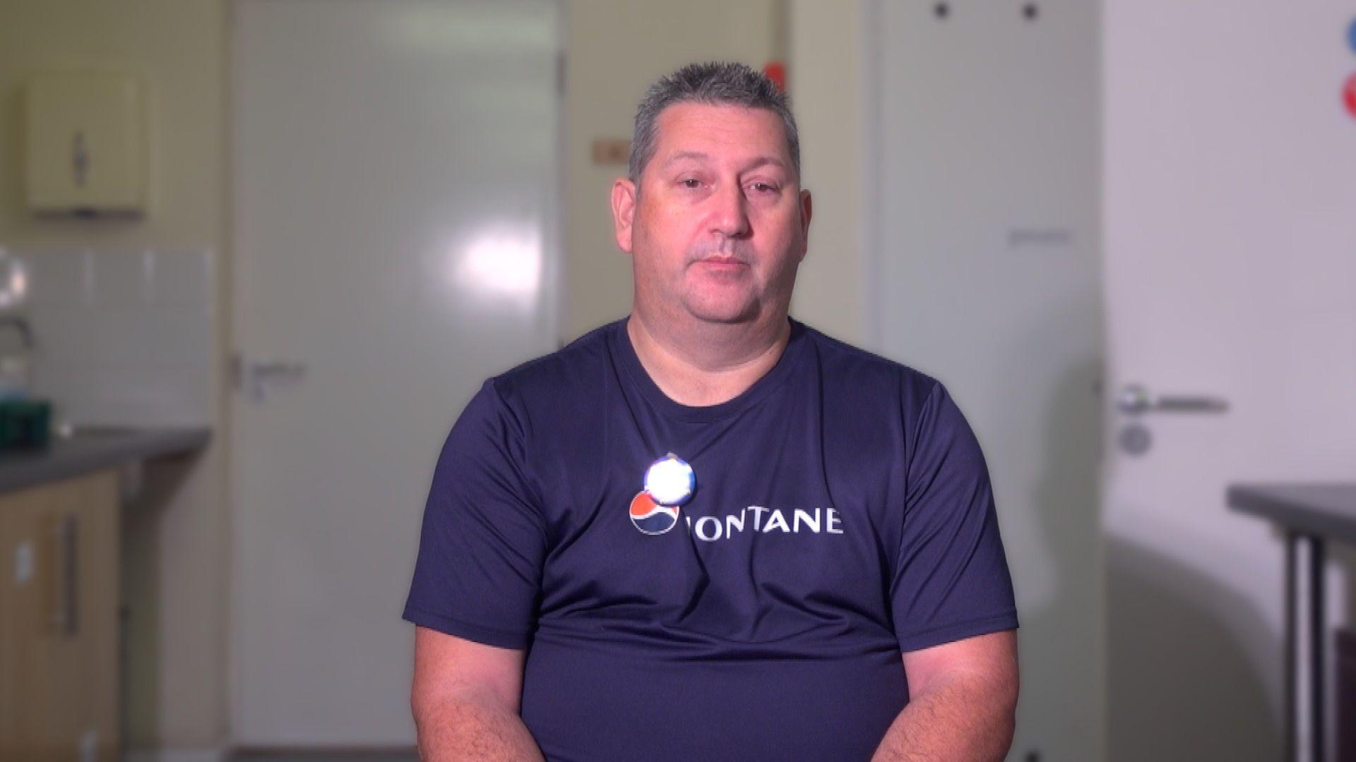 A man with short grey hair is facing the camera. He is wearing a navy t-shirt with white writing on it which can't quite be read. Behind him are white doors