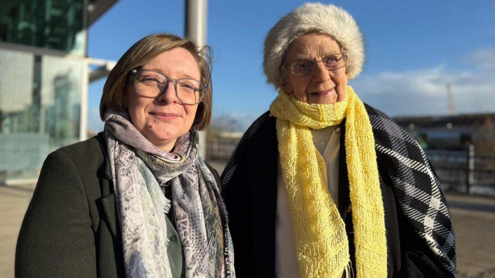 Former shipyard worker Catherine Jamieson stands on the right wearing a furry hat and a yellow scarf, with her granddaughter Angela Stevenson standing next to her wearing a black coat and a patterned scarf.