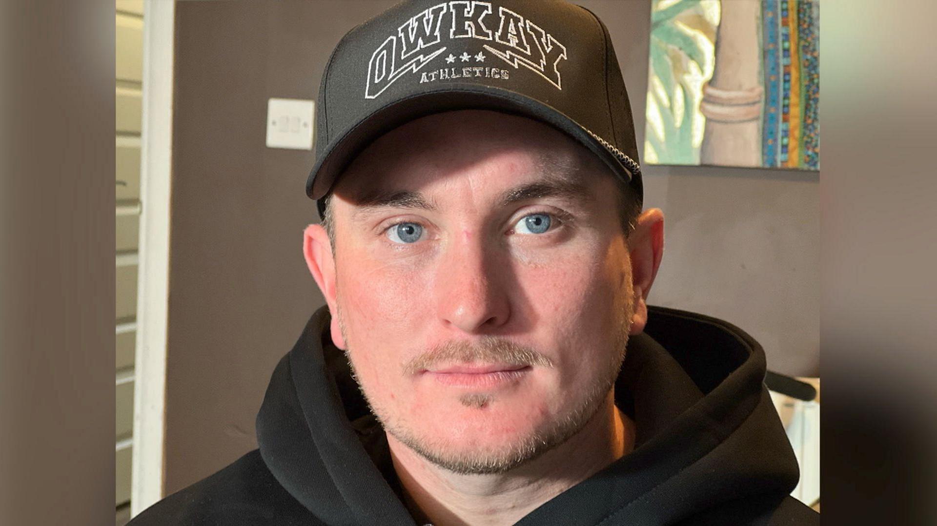 A man with a stubbly mustache and beard looks down the lens with a neutral expression. He's wearing a black cap and black hoodie.