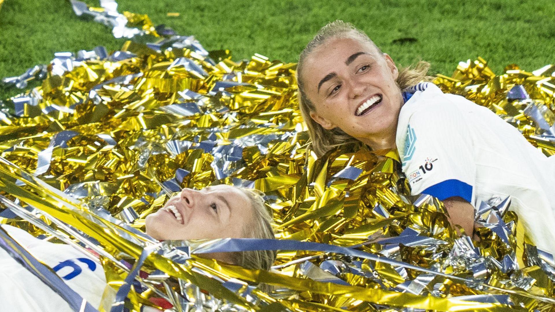 leah williamson lies down in gold and silver confetti looking delighted while georgia stanway looks off to the side while smiling