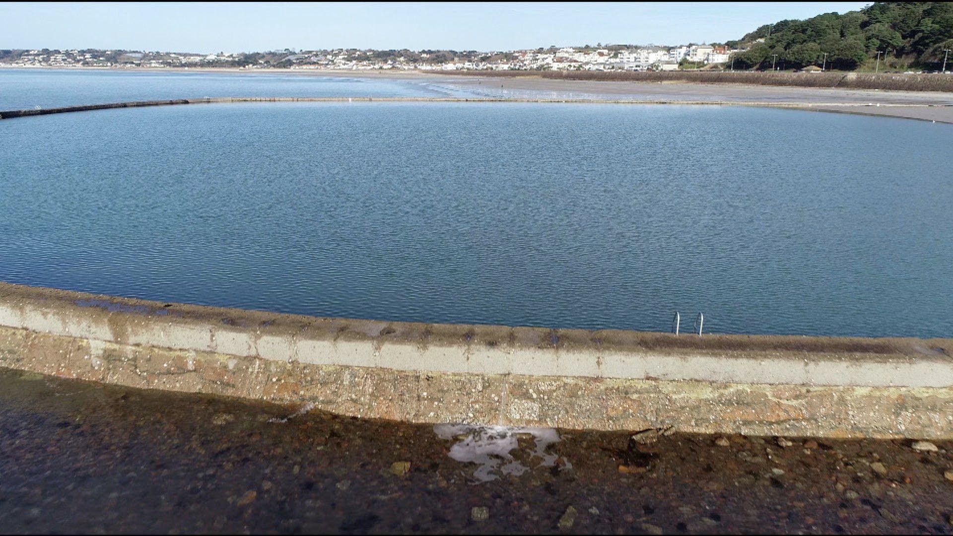 A drone shot of the Victoria bathing pool, it's a circular bathing pool with Saint Aubin's bay in the background