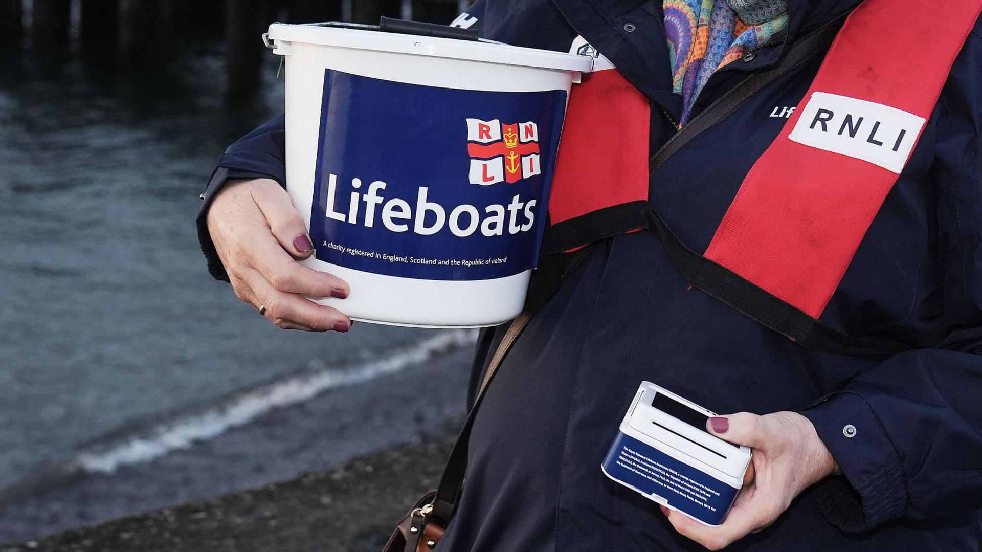 A person holding a charity bucket with blue RNLI lifeboats branding. The person is wearing a lifejacket and is holding a card payment machine. They are standing by the seaside. 