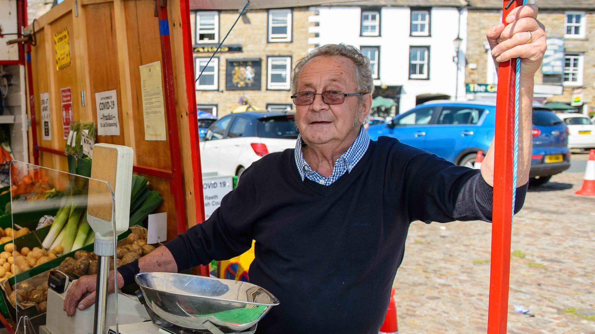 A 76-year-old man in a blue jumper and checked white and blue shirt stands at a market stall next to a weighing scale and a selection of vegetables