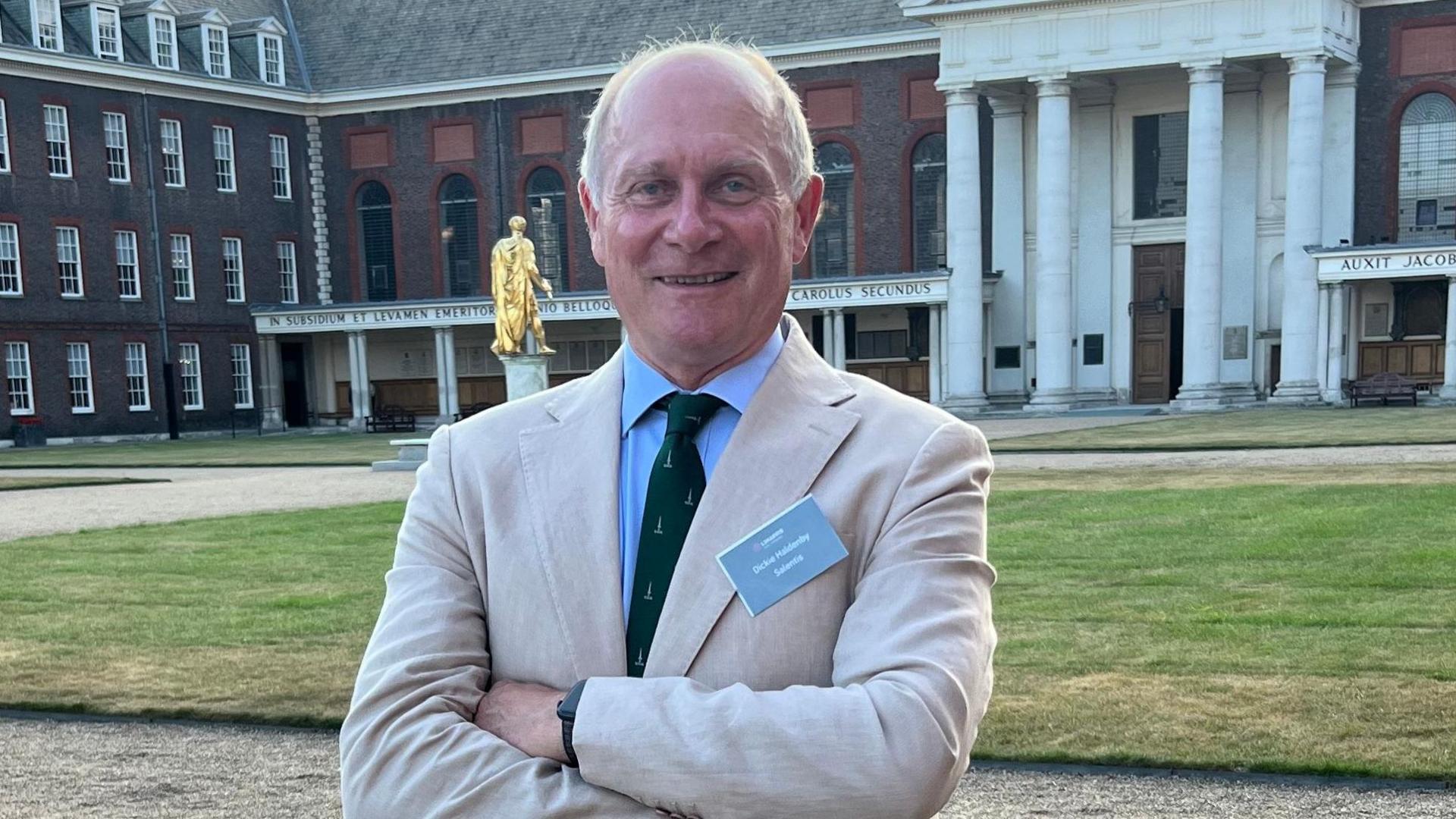 Richard Haldenby is standing with his arms crossed wearing a cream jacket and a blue shirt and green tie, arms crossed, in an open courtyard. He has a name badge on his jacket.