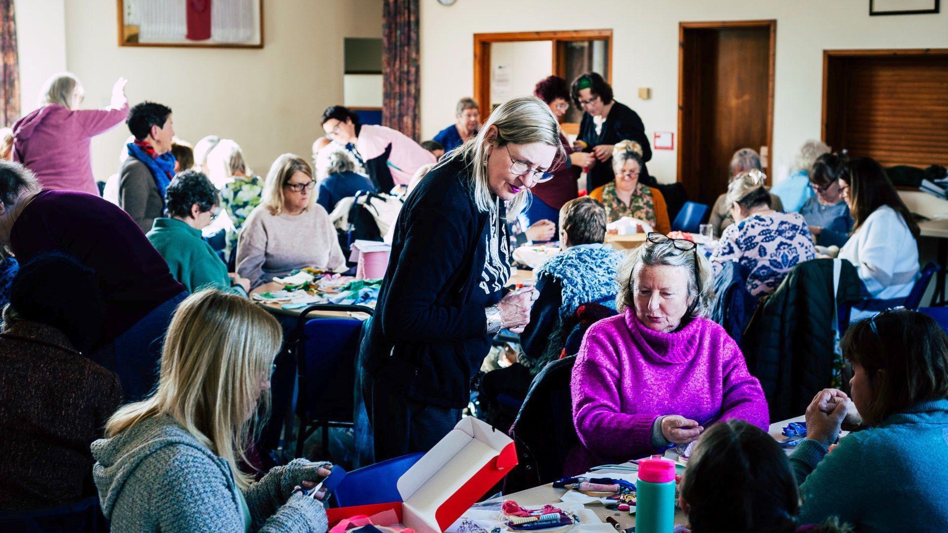 A wide shot of women gathered in a large hall, all sat at tables sewing and chatting. In the foreground, a woman with long blonde hair wearing glasses, a black and cream striped blouse and black cardigna over the top is looking down at a lady's work.