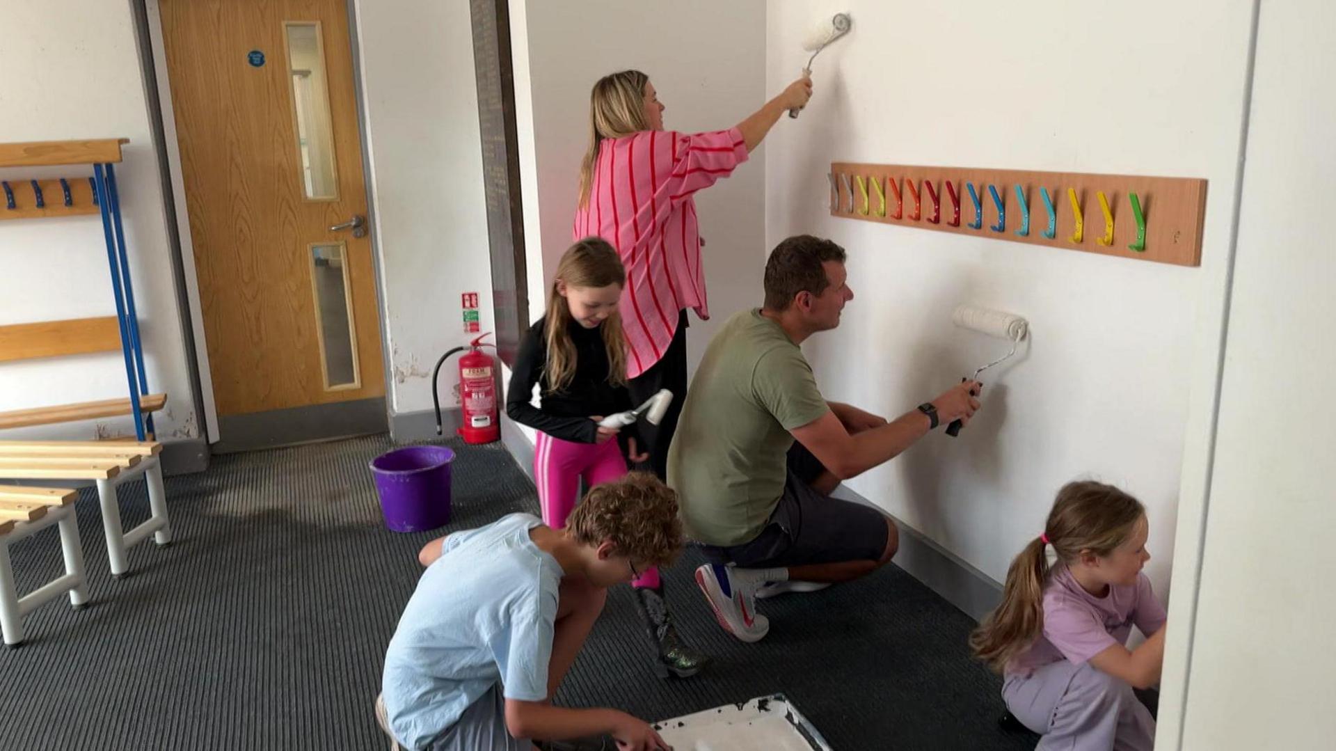 Two adults and three children are all painting the same wall in a school corridor. Colourful coat hooks are in the middle of the wall and small wooden benches can be seen on the left.