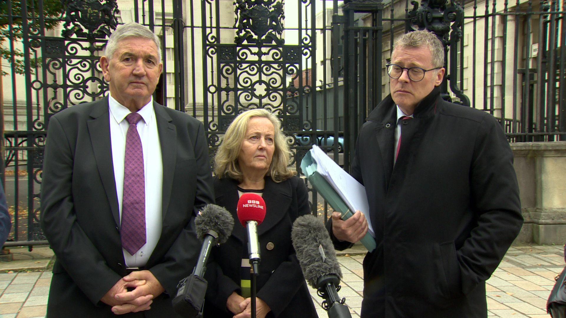 Peter Jackson, with grey hair, in grey suit, with white shirt and purple tie, Gay Jackson in dark coat, with blonde hair and Kevin Winters with grey hair, glasses and long dark coat, outside the High Court in Belfast.