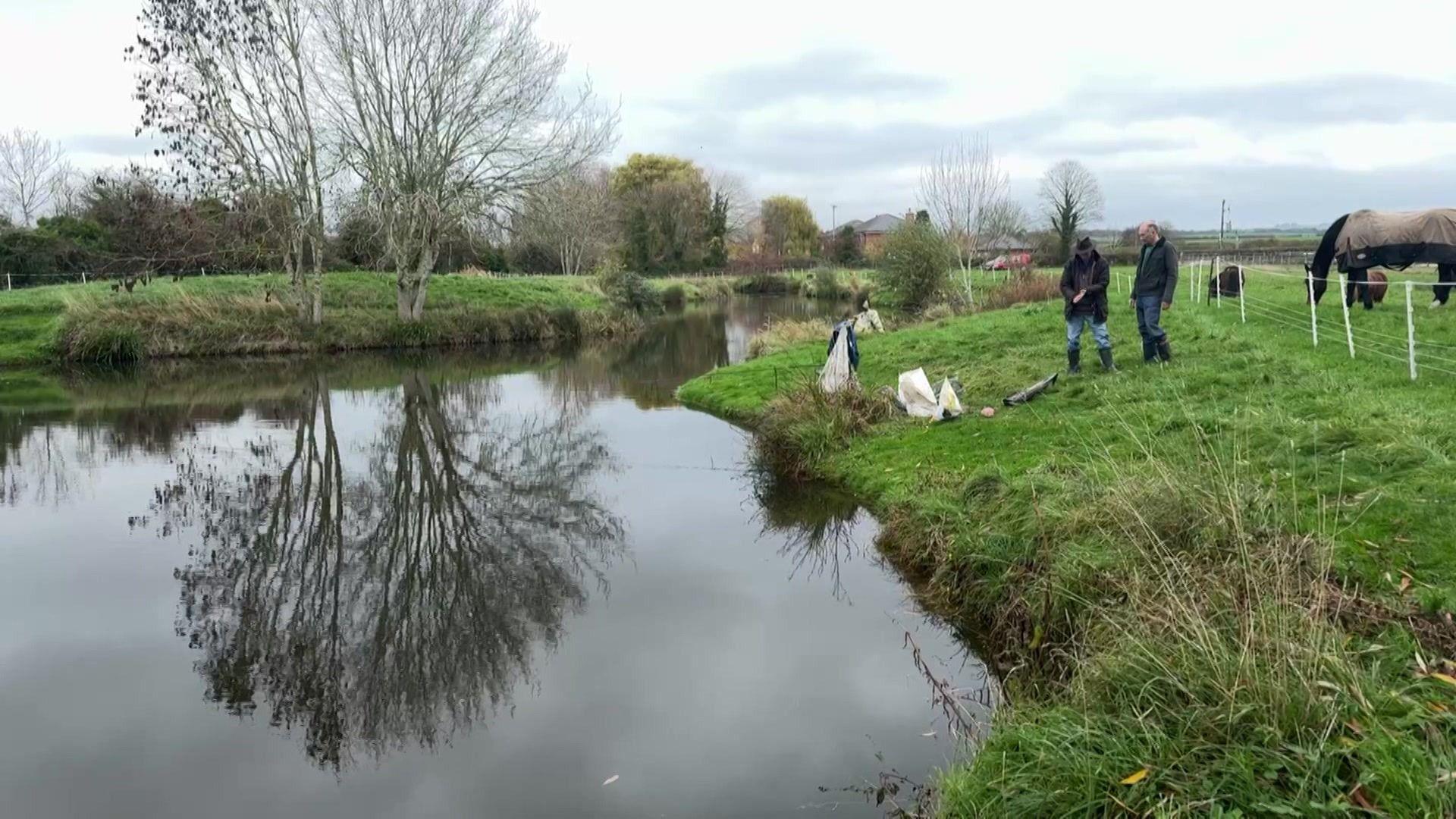 A wide shot of a small man-made fishing lake, surrounded on all sides by grassy banks. Two men can be seen standing near the right-hand bank. Trees are in the background