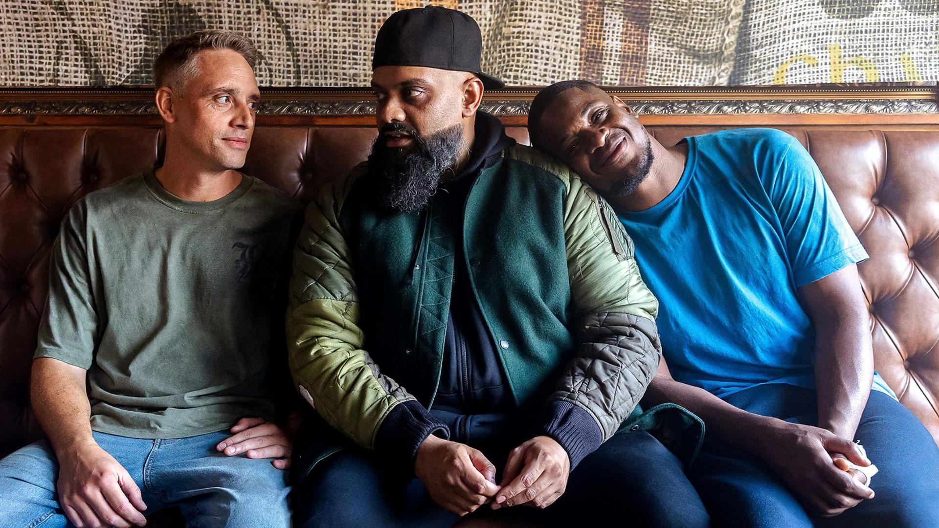 Three characters, Harper (Perry Fitzpatrick), Mobeen (Guz Khan), Nate (Tolu Ogunmefun), sit on a sofa. Two are talking to each other and one has his head on Mr Khan's shoulder, smiling.
