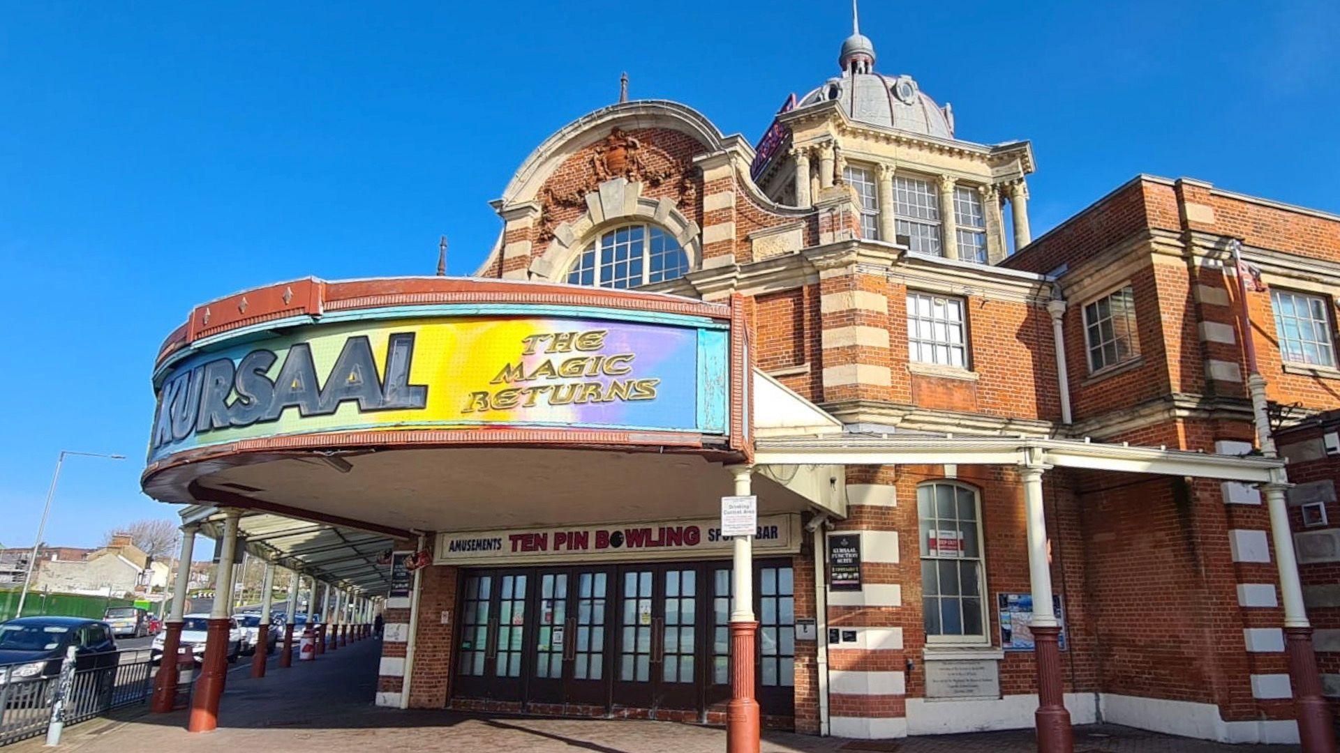 The Kursaal building in Southend-on-Sea. It has a brick front with a large tower and domed roof. There is a colourful curved sign at the front which says "Kursaal - the magic returns"