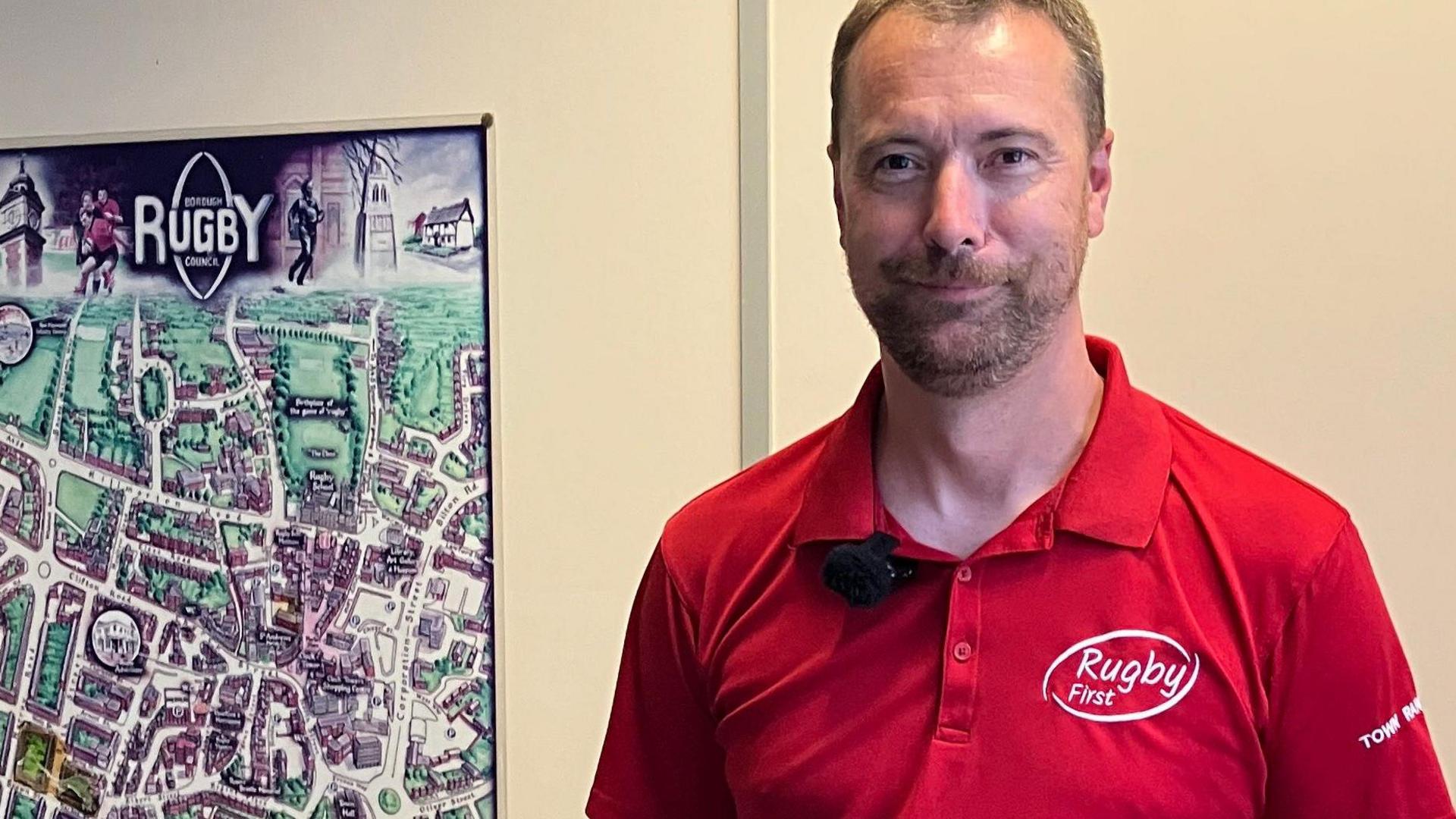 Rob Hallam stands next to a map of Rugby town centre. He is wearing a red top which is part of the rangers' uniform. The polo shirt has the Rugby First Logo on it in white, and the words Town Ranger are on the sleeve.