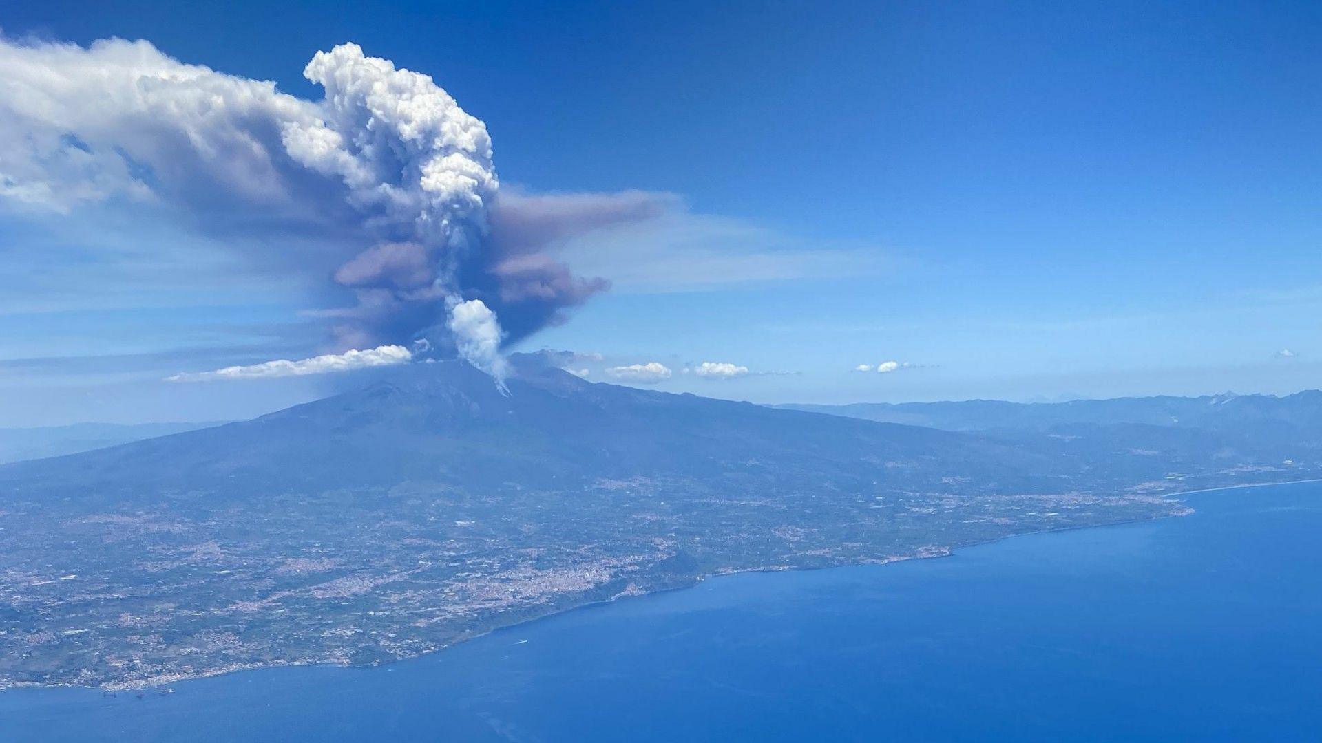 A picture of Mount Etna erupting from a plane. There is grey and black cloud coming out of the top of the volcano. The sky is blue. The ground is green and grey. The sea is a darker blue than the sky.