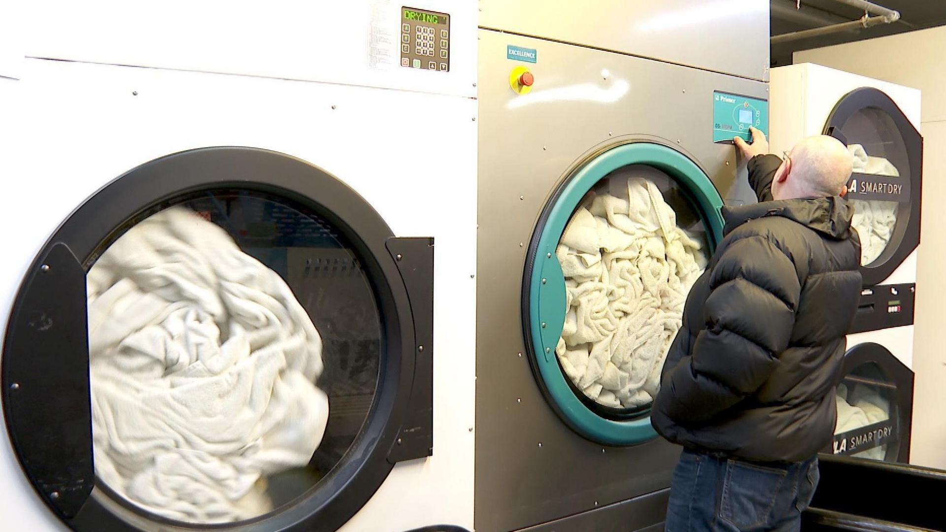 Mr Mason, who is wearing a black puffer-style jacket, stands at the controls of a large washing machine. There are white sheets in the machine, and ones next to it.