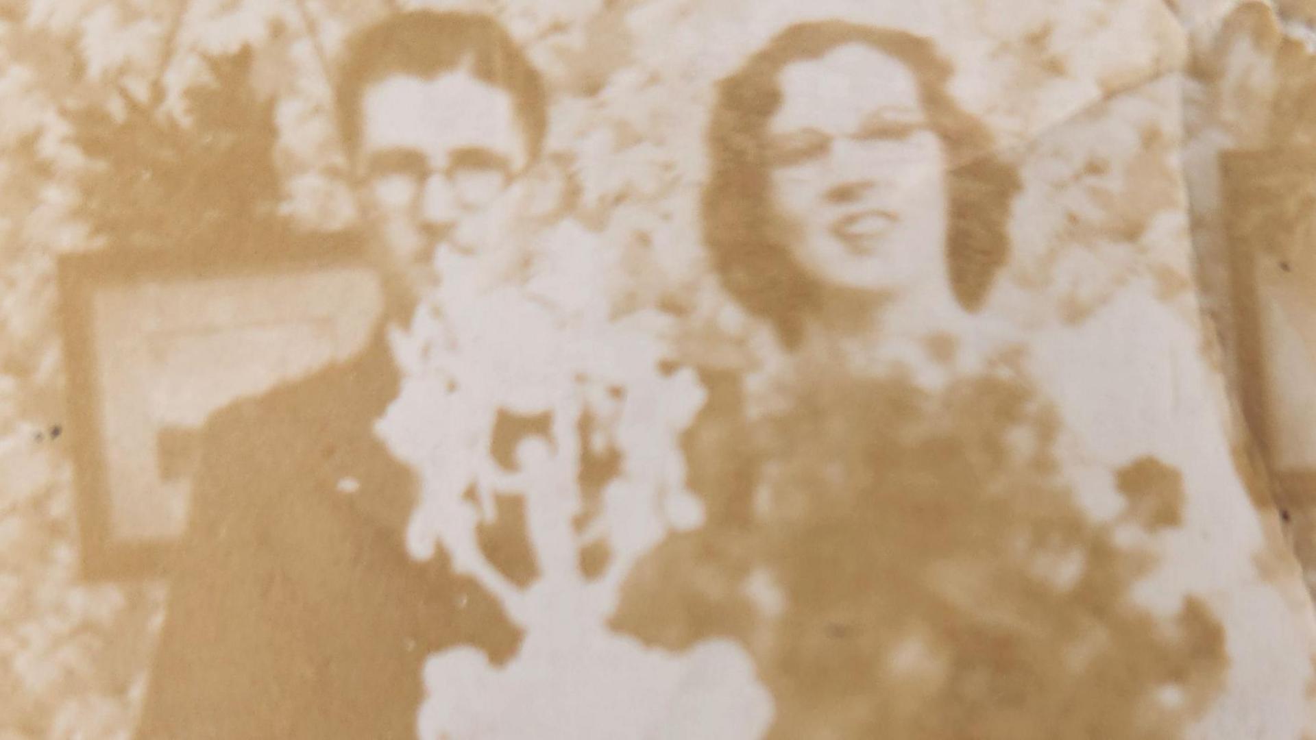 A sepia image of a couple standing in front of a tiered wedding cake. The man is wearing a dark suit and the woman a wedding dress.
Floral wallpaper decorates the wall behind.