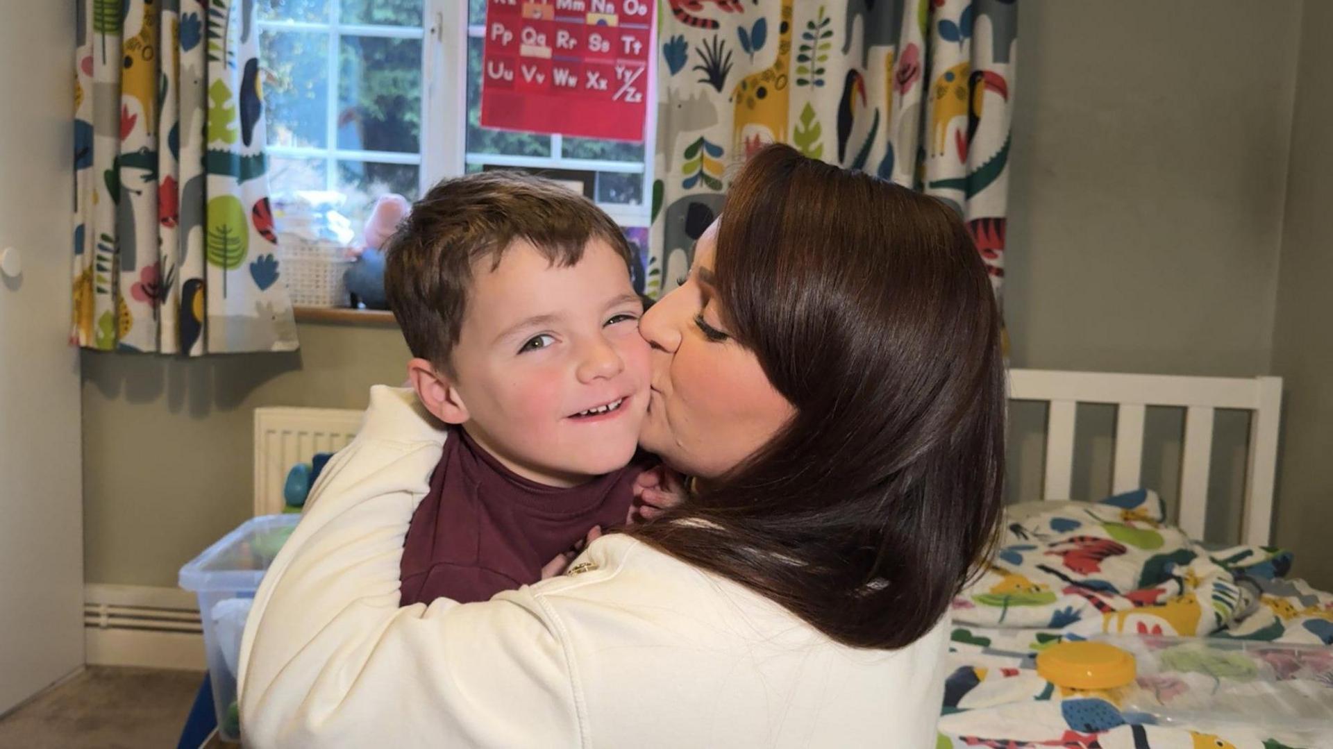 Max looks at the camera and is being held by his mother Nirvana who is kissing him on the cheek. They are in a child's bedroom with colourful animal curtains in the background and a matching duvet cover also visible on the bed.