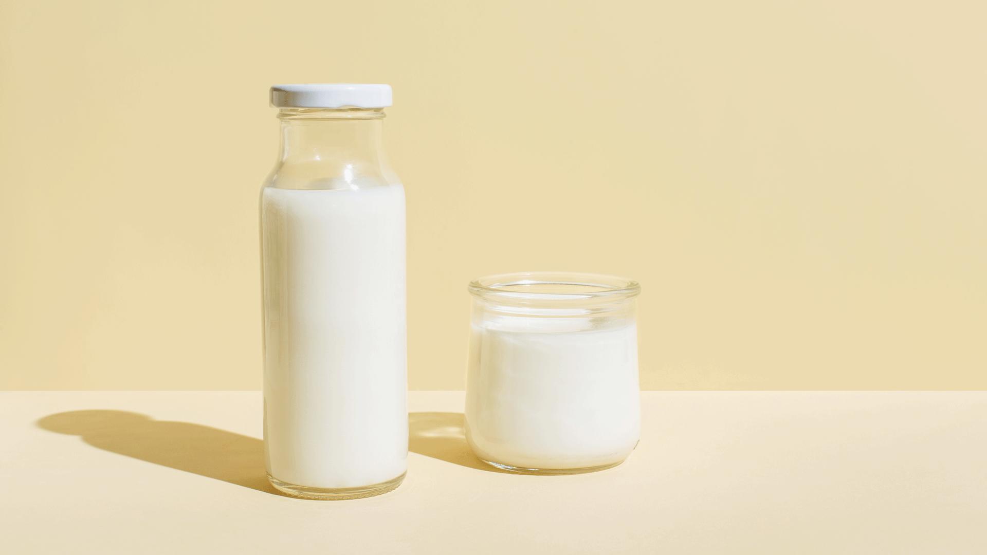 A glass bottle of milk and small glass of milk next to it. With a pale yellow background.