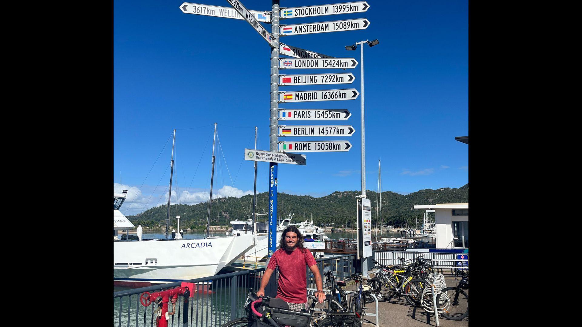 A man in a red t-shirt standing with his bike by a waterfront in Queensland, Australia.