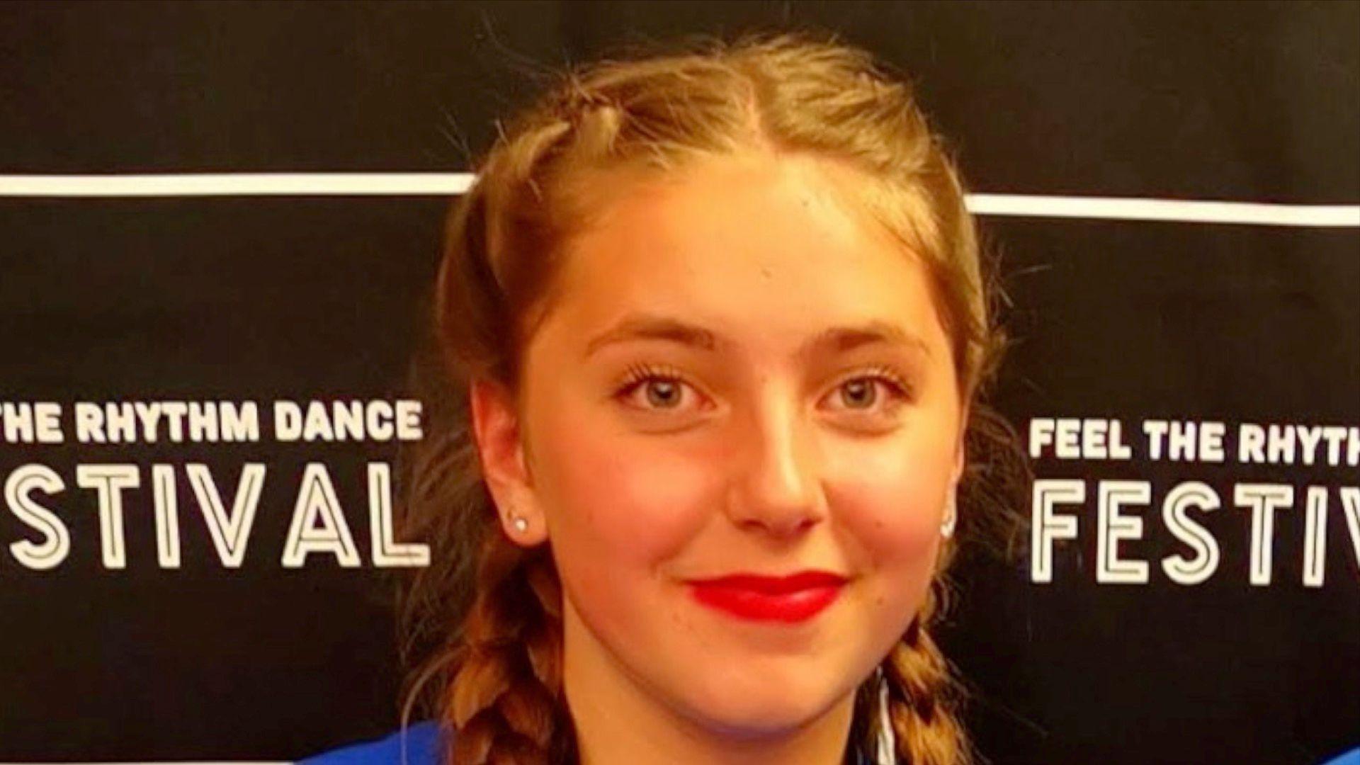 Chloe Longster smiles at the camera. She is wearing pigtails in her hair with stud earrings in and red lipstick. The picture is taken at a dance festival where she was awarded a certificate.