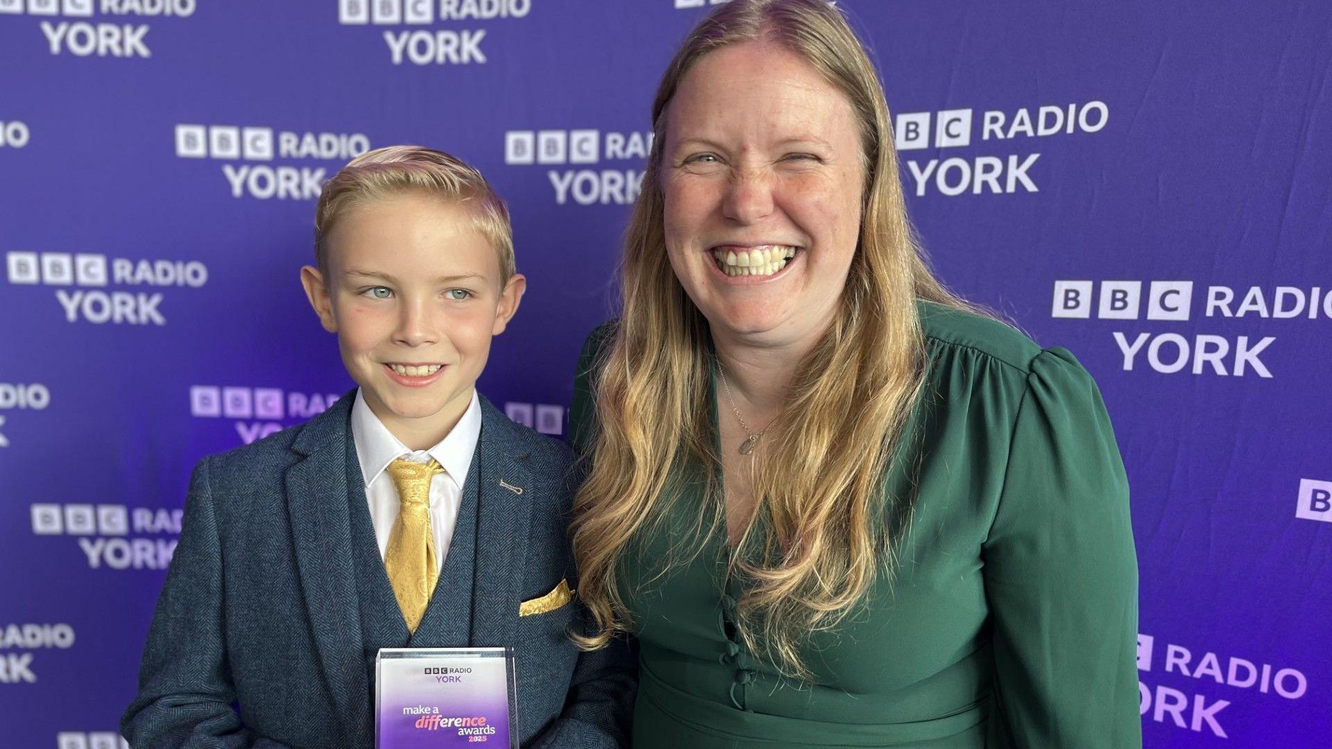 A young boy stands next to his mum. The boy has blue eyes, blonde hair and is wearing a smart blue-green tweed suit with a waistcoat and yellow tie. He is smiling and holding a glass award with 'Make A Difference Awards 2025' on it. His mum is wearing a green dress and has long blonde hair and is wearing a silver pendant necklace. She is also smiling. Behind them is a purple backdrop with 'BBC Radio York' on it. 