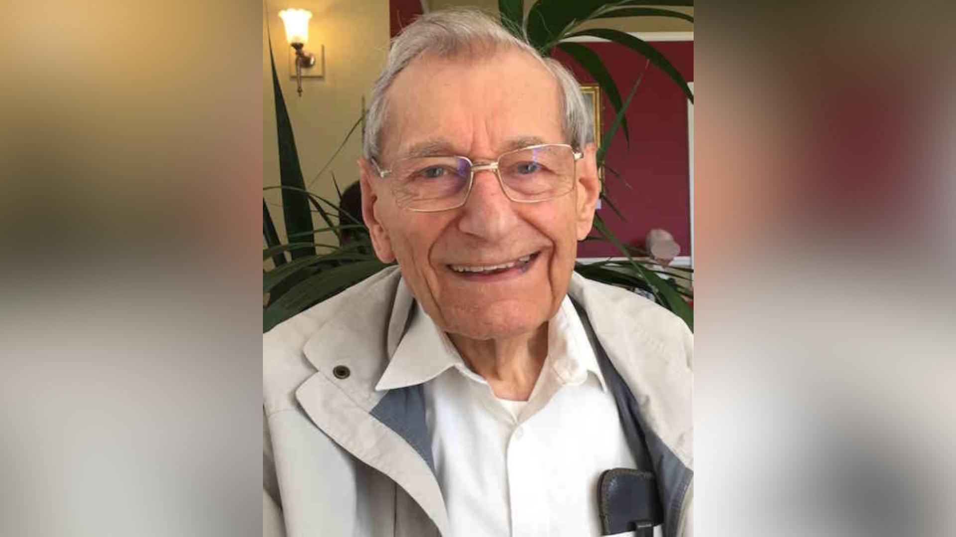 Mike Gibson smiles widely at the camera. He has thin grey hair, wears glasses and a light grey coat with a white shirt underneath.