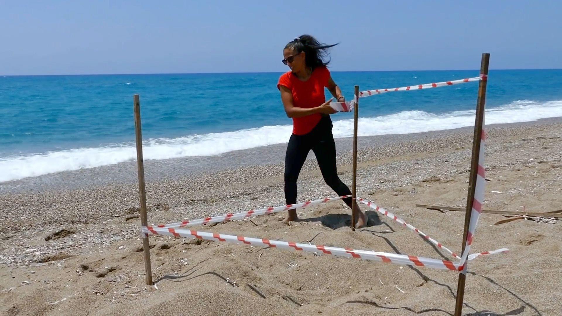 A woman on a beech is wrapping plastic tape around three wooden poles which are arranged in a triangular shape, behind her the sea is visible