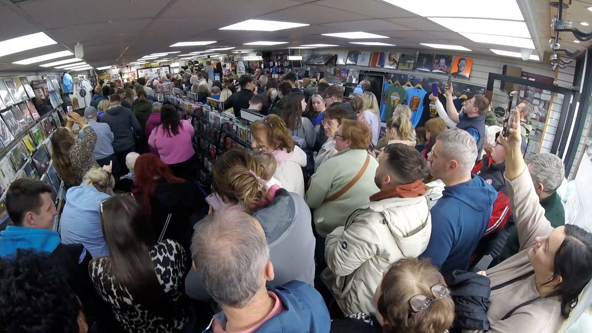 A wide shot showing the inside of Stewart's which is completely full of people crowding around to see Daniel O'Donnell. Many peole are holding up phones to film O'Donnell, who can barely be seen in the distance.