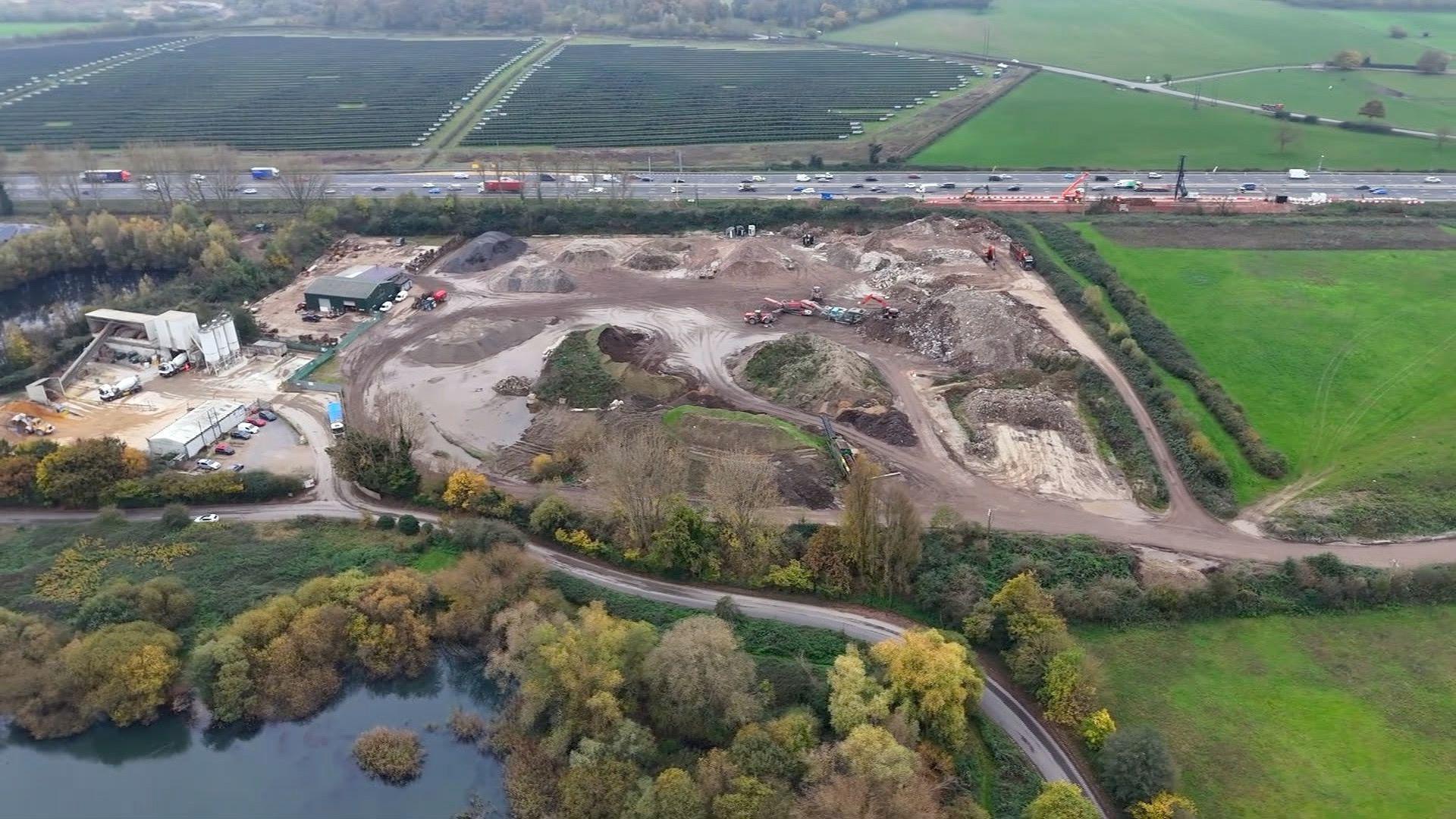 A drone shot of the incinerator site shows a section of construction, with a road on the side, trees and fields surrounding the area and a section of water