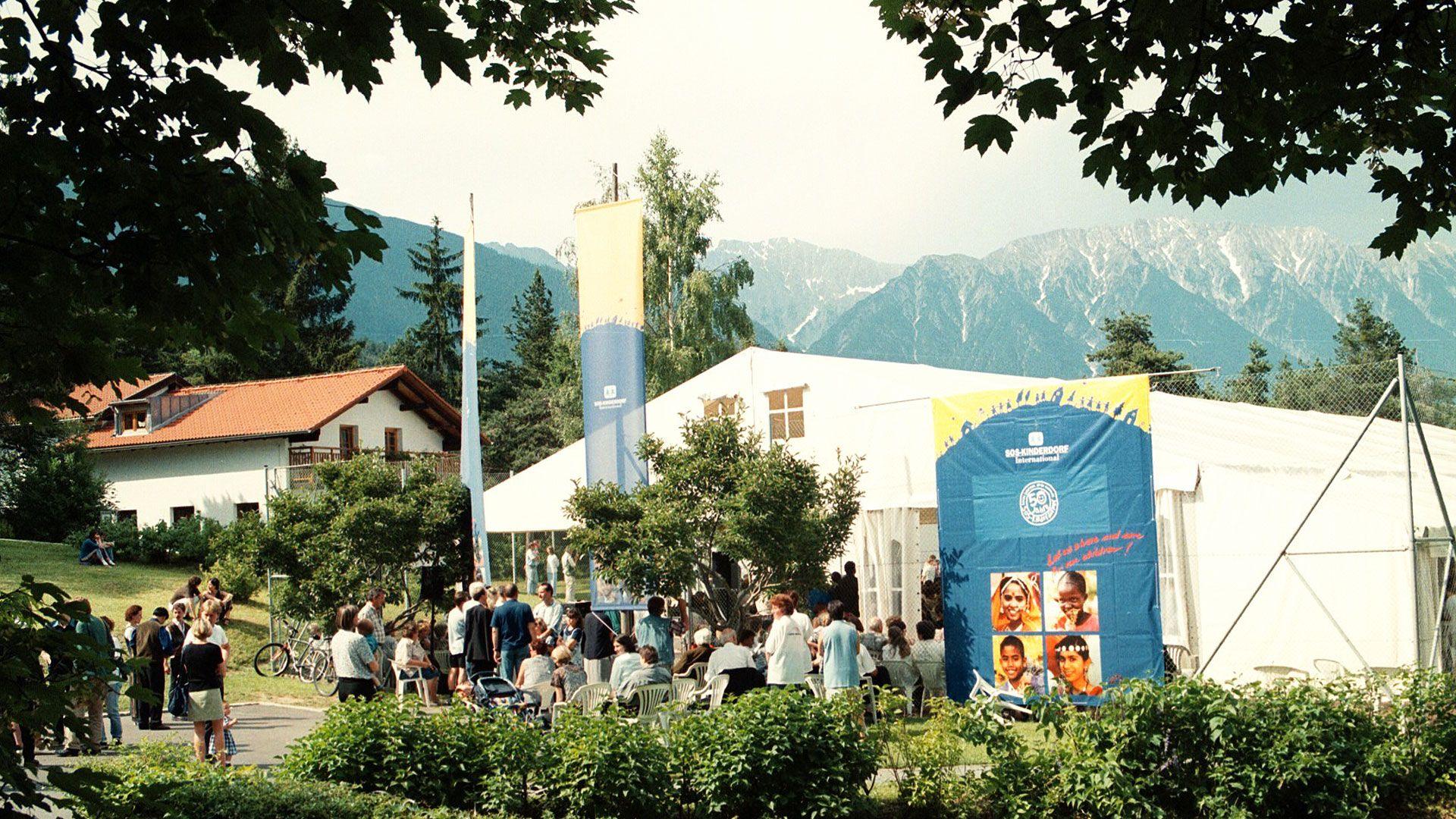 A small crowd mills in front of a marquee and a poster with mountains in the background