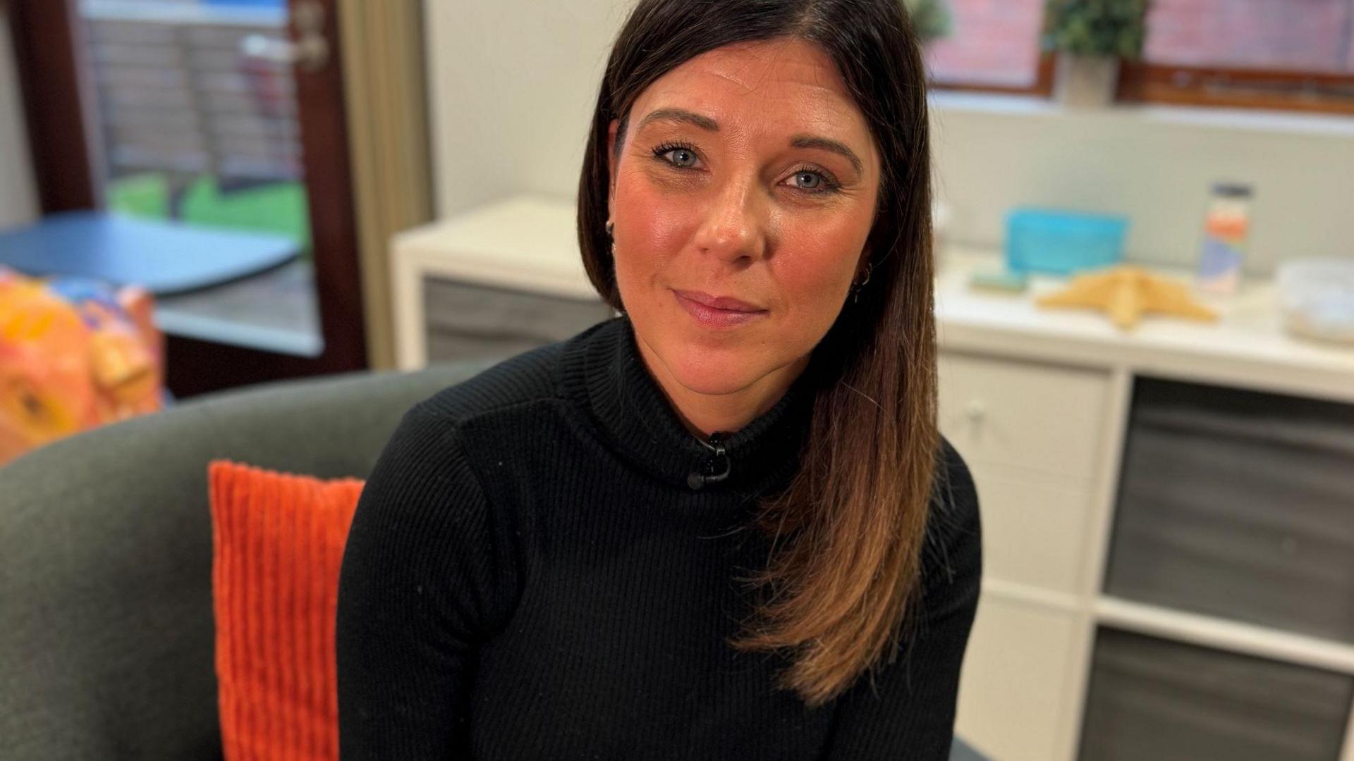 Elinor Eustace looking at the camera. She has long dark hair and is wearing a black jumper and sitting on a sofa with an orange cushion behind her. 