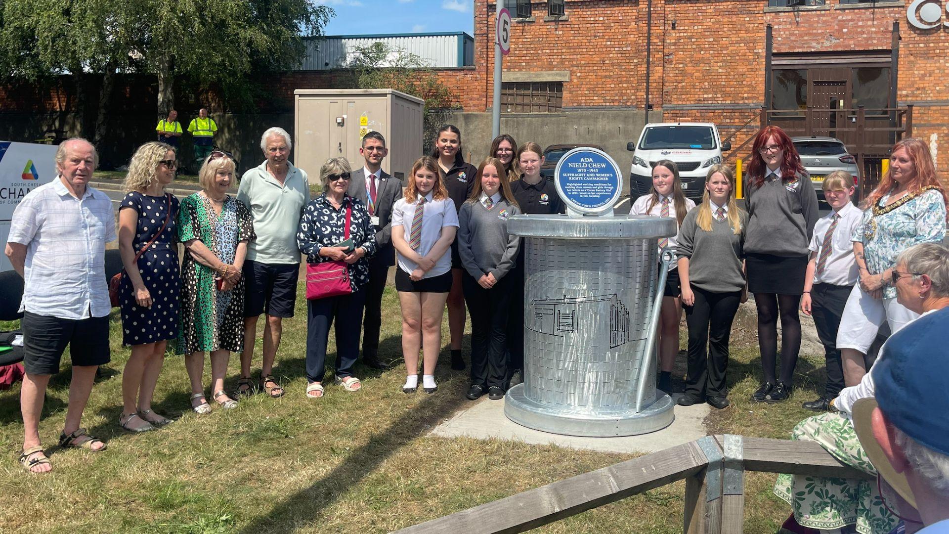 A blue plaque stands on a metal plinth moulded into the shape of a large cotton bobbin and needle, decorated by a laser cut outline of a factory. To the left of the plinth stands two men and three women dressed in summer clothing. Behind the plinth stand nine females dressed in college uniform. A woman wearing mayoral chains stands to the right. The plaque and the crowd stand on a grassy area in front of a brick building.
