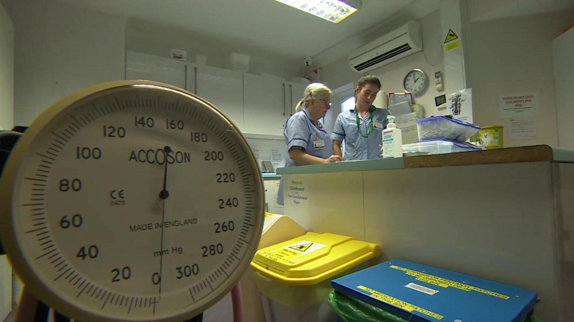 Two nurses stand in a room with medical supplies, there is a large set of scales in the forground which has a circular face and cream outer frame. There are medical waste bins with bright yellow, blue and cream lids and the nurses stand over a worktop looking at something. The nurse on the left has white hair tied back in a ponytail and wears clear framed glasses and a pale blue nurses uniform. The nurse to the right is slightly taller and has dark brown hair tied back in a bun.