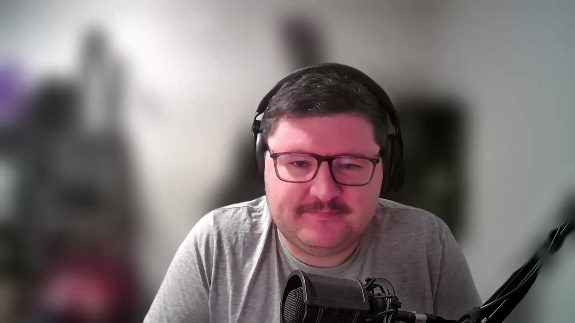 A still taken from a zoom call, featuring Mr Brown, who has short dark hair, dark rimmed glasses, and a dark moustache. He talks into a microphone and wears headphones.