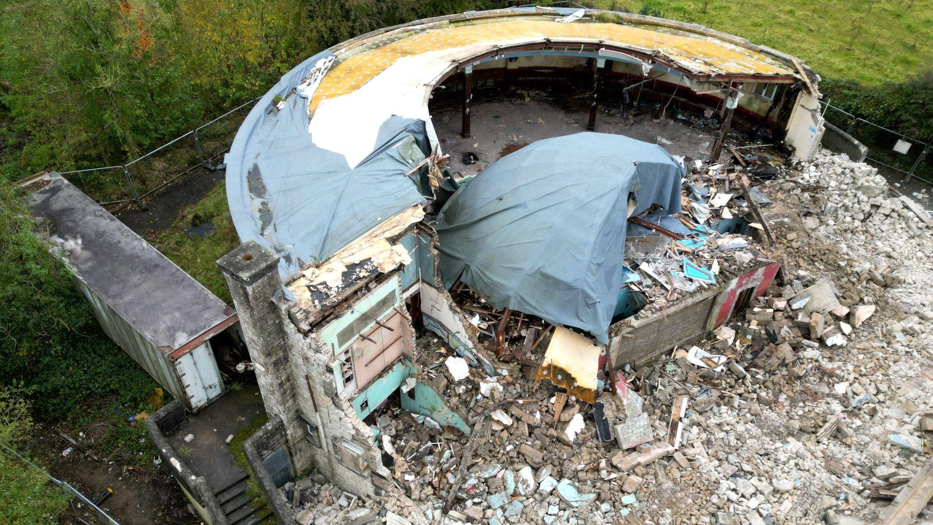 Images of Edendork Hall with a lot of brown rubble on the floor and a frame of the building with the back wall still intact. The dome is mostly destroyed, half of its shape remains and it's covered with a blue cover. Behind the demolished building is a green field. 