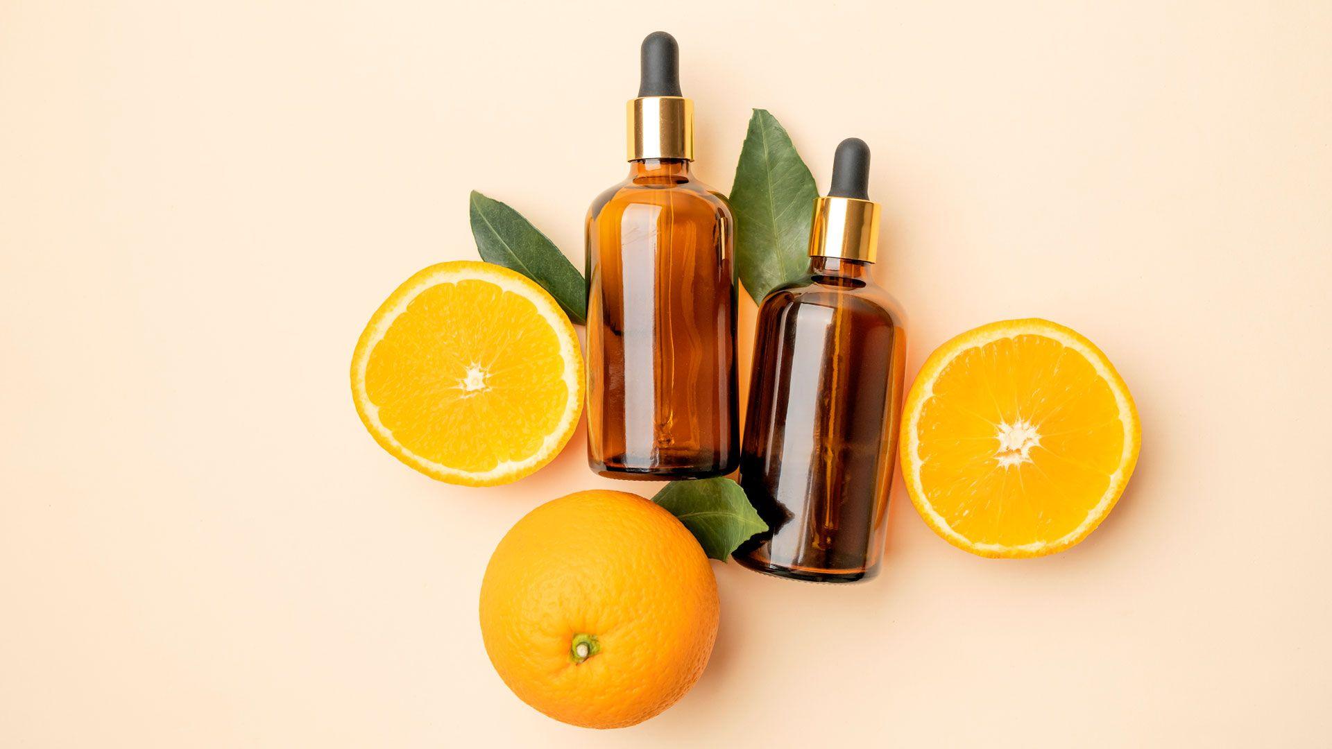 Two slices of oranges and one whole orange and placed around two brown bottles with pipettes in them. Suggestion is that the bottles contain vitamin c face serum which is frequently used to help skin improve. 