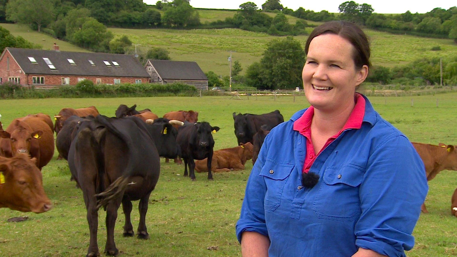 A farmer - a woman in her 40s wearing a blue shirt with rolled up sleeves - stands in a field surrounded by calves on a gloomy day. A farmhouse can be seen in the distance.