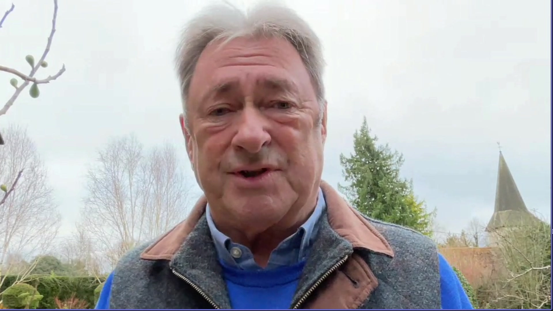 Alan Titchmarsh, is standing in his garden and wearing a blue jumper, blue shirt and grey gilet.