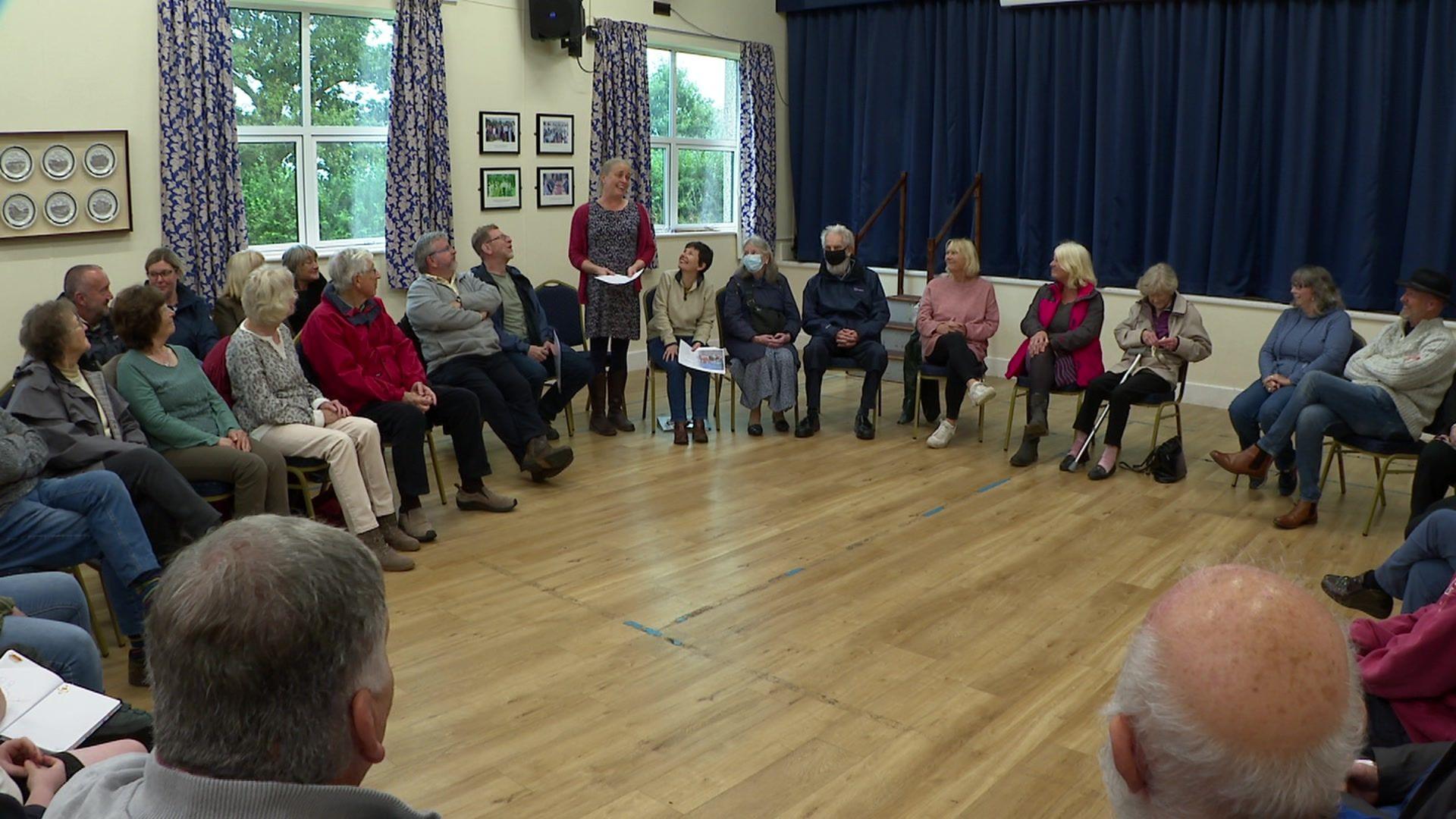 A hall with many people in the room listening to a woman talking to them about the plans for the shop