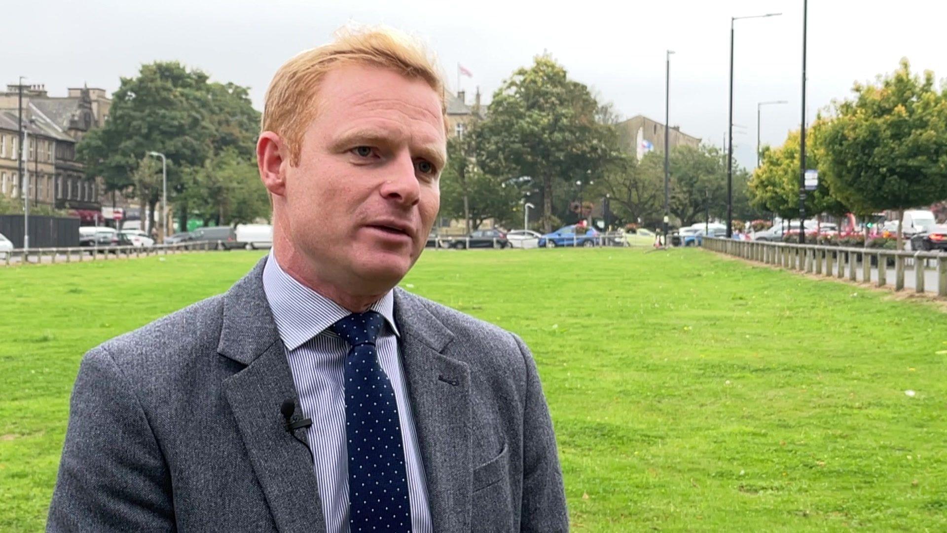 A photo of Robbie Moore MP, stood by a green space near roads and buildings in his constituency. 