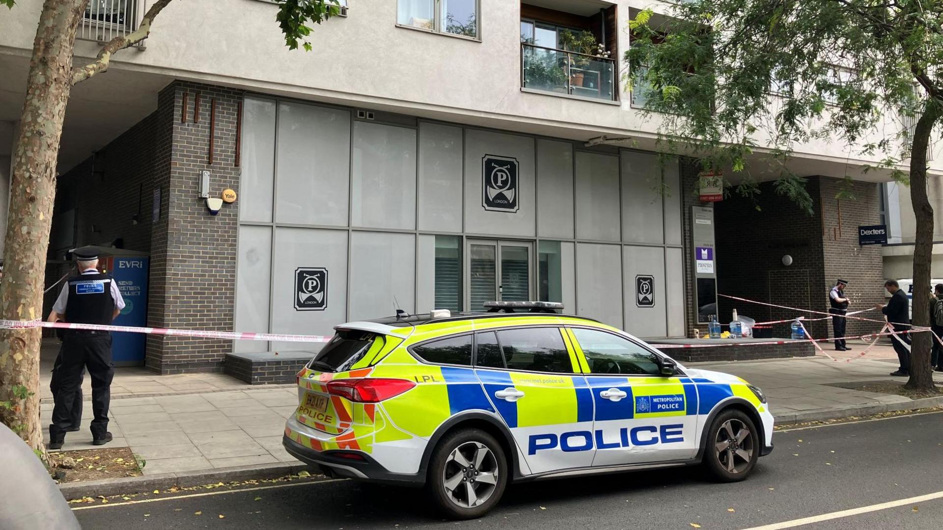 A police car parked outside a grey building and several police officers nearby behind police tape