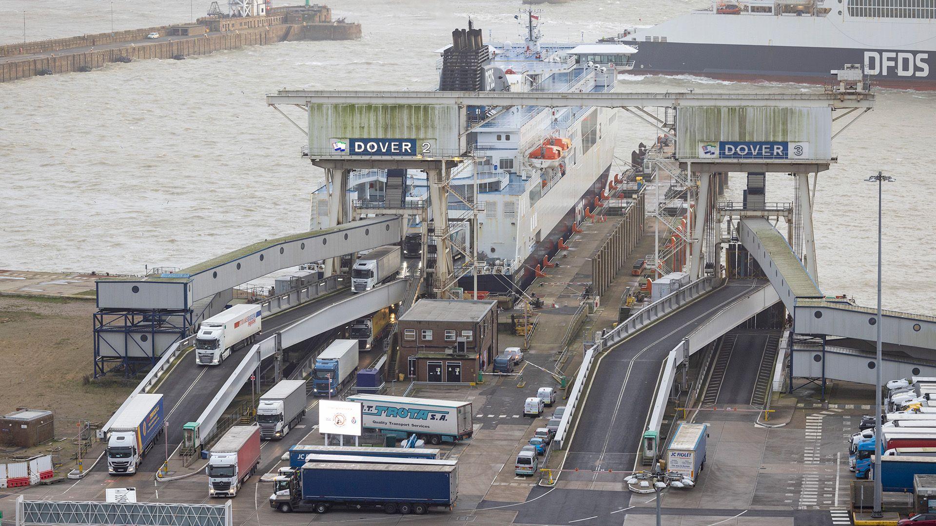 A port terminal where numerous trucks and trailers are queued on ramps leading to a docked ferry. The weather is grey and overcast.